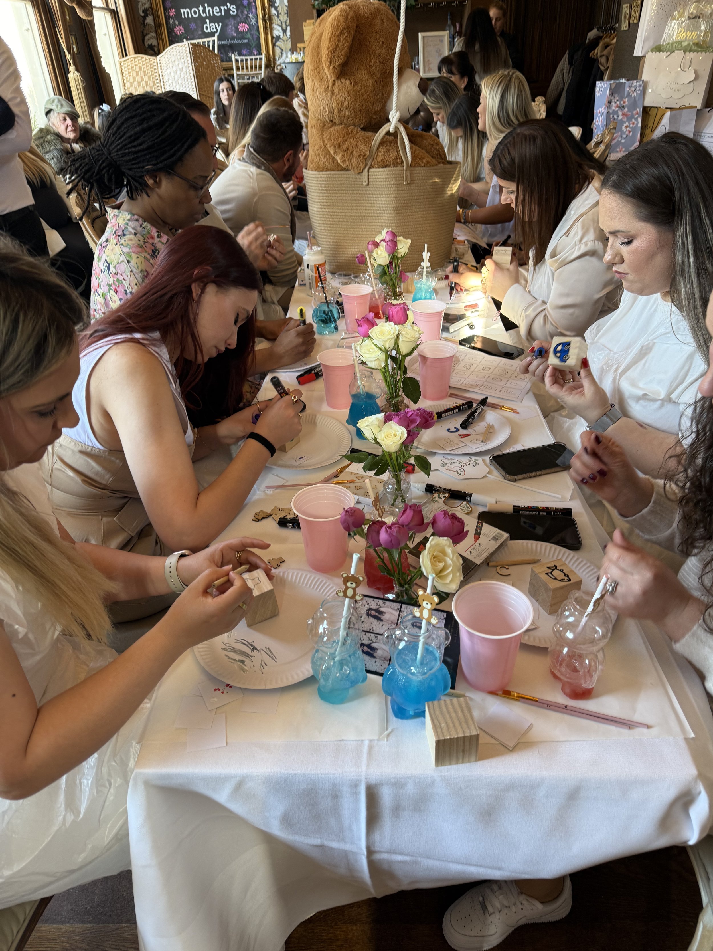 Group of women gathered around a table at a mother's day celebration, making crafts with rubber stamps, surrounded by flowers, drinks, and party decorations.