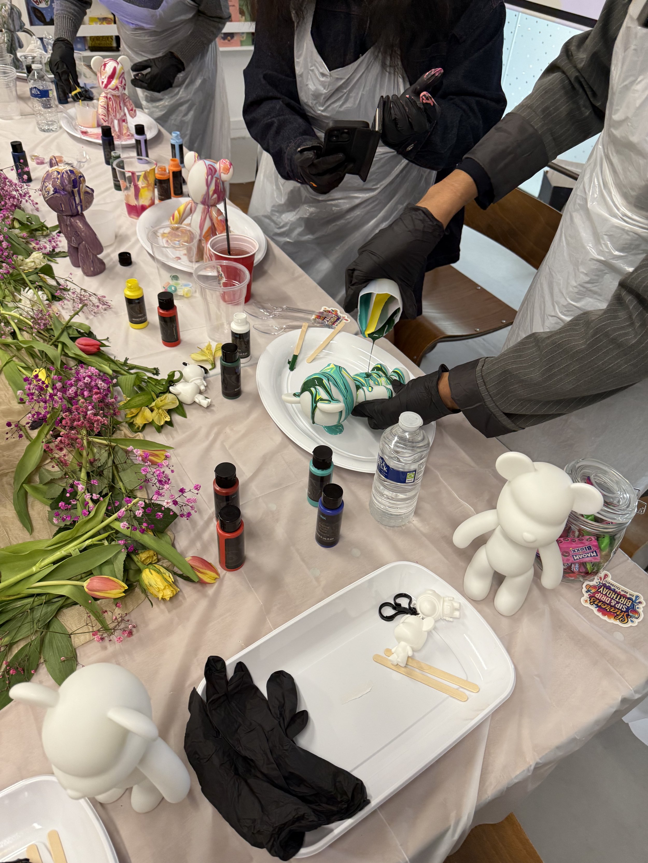 People decorating bear-shaped soap or figurines with colorful swirl designs at a crafting table surrounded by paints, brushes, flowers, and craft supplies.