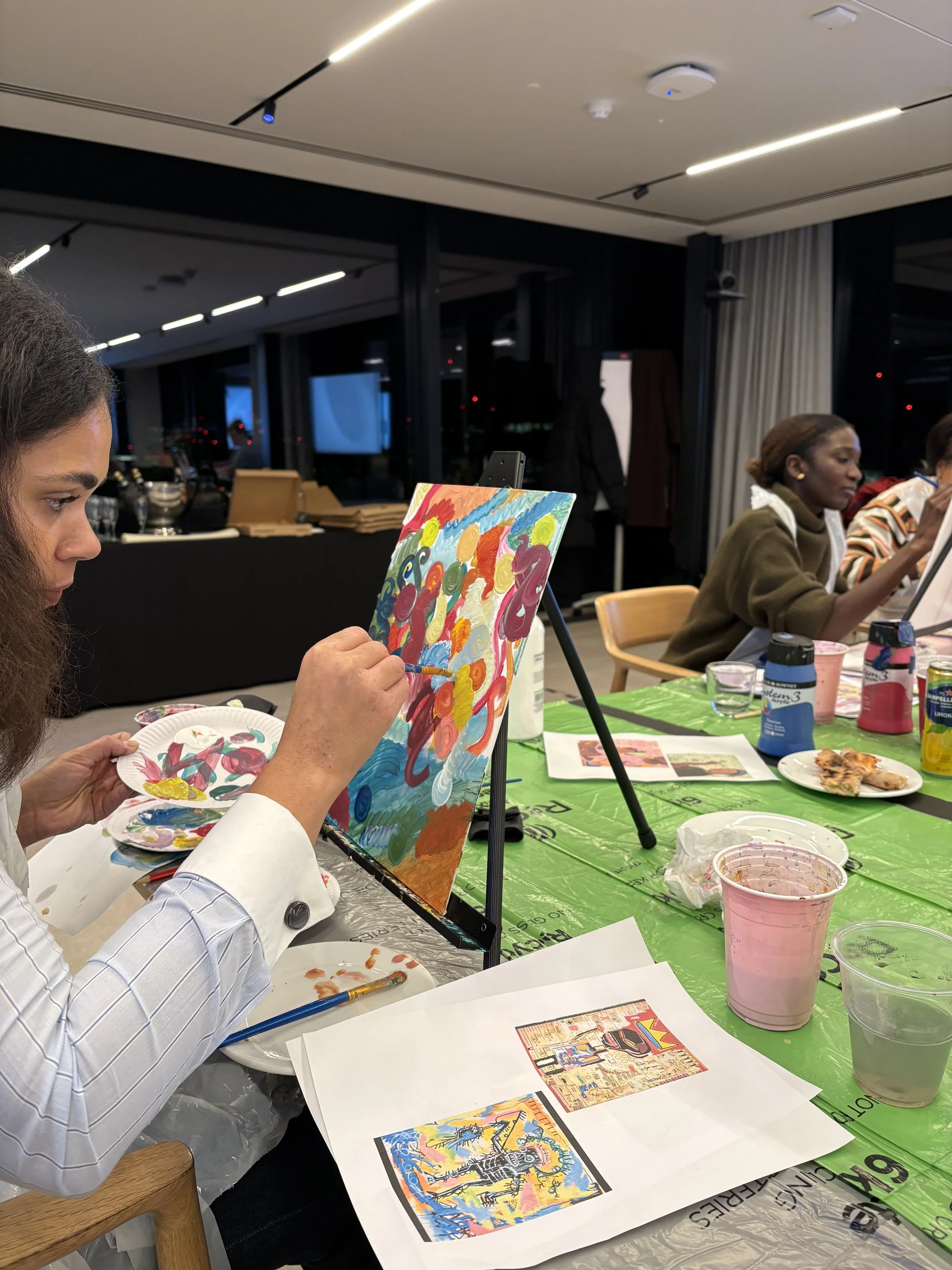 Woman painting an abstract colorful artwork on a small canvas at a table during an art event, with other participants and art supplies visible.