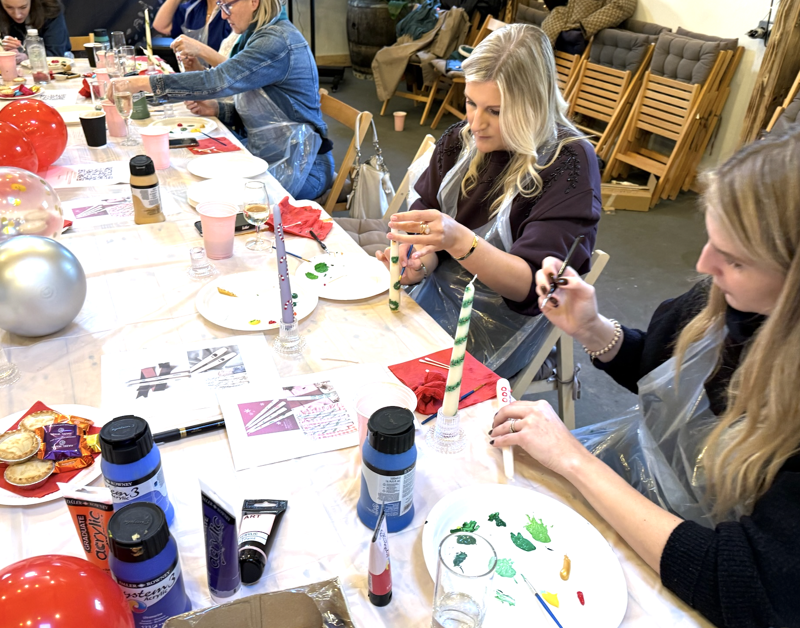 Women sitting at a table painting and decorating candles during a craft party, with Christmas-themed decorations and supplies around them.