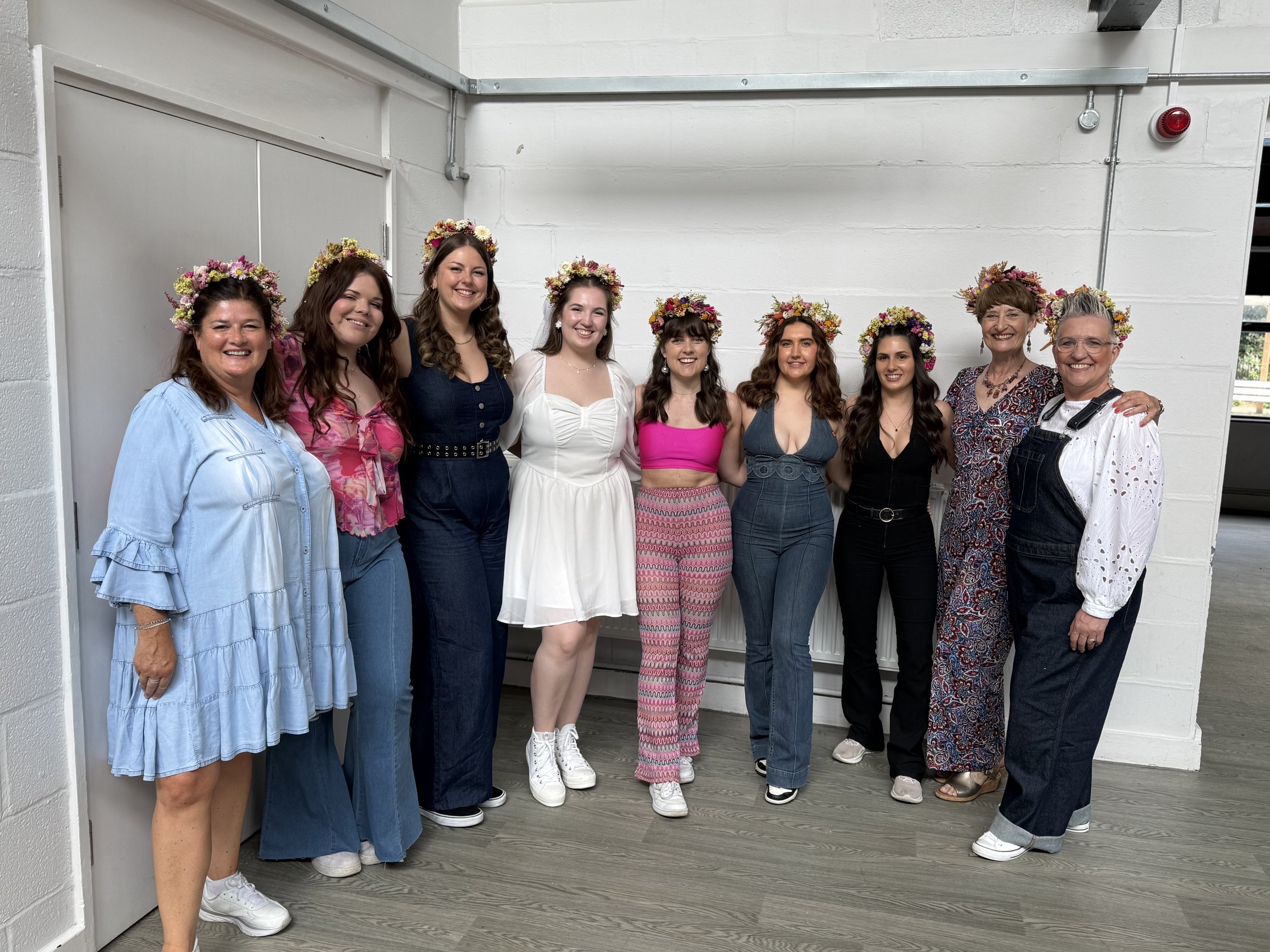 Group of women and girls, some wearing flower crowns, standing in a line indoors with white walls and a gray floor, smiling for the camera.