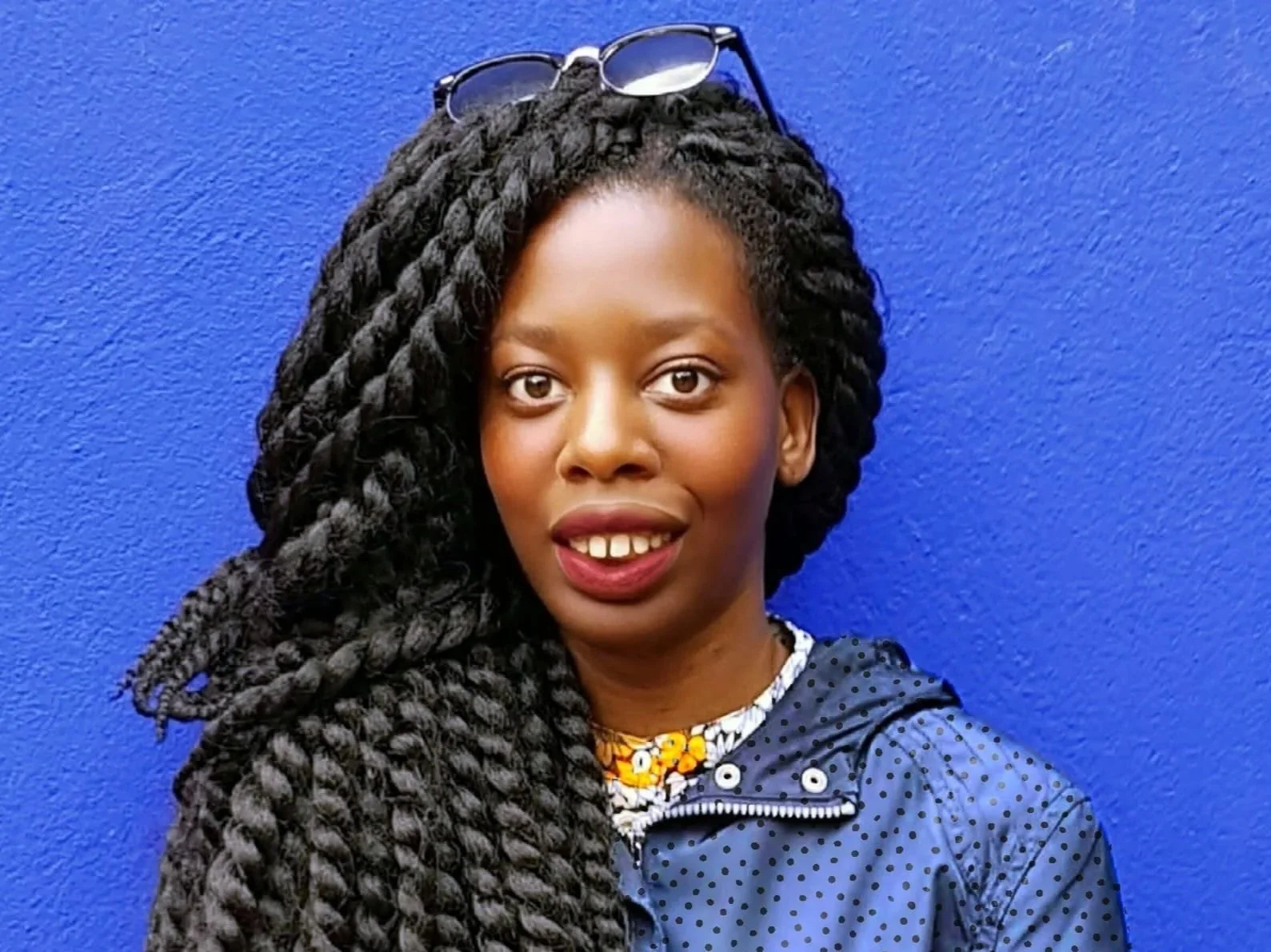 A woman with dark skin and long, thick, twisted black hair stands in front of a blue wall. She is smiling, revealing slightly uneven teeth, and wears glasses on her head, a blue polka dot jacket, and a yellow and white floral necklace.