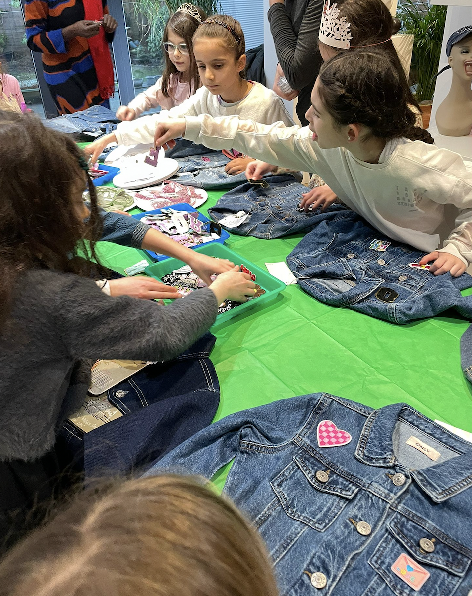 Children and adults gathered around a table with denim jackets decorated with patches, stickers, and pins, engaged in a crafting activity.