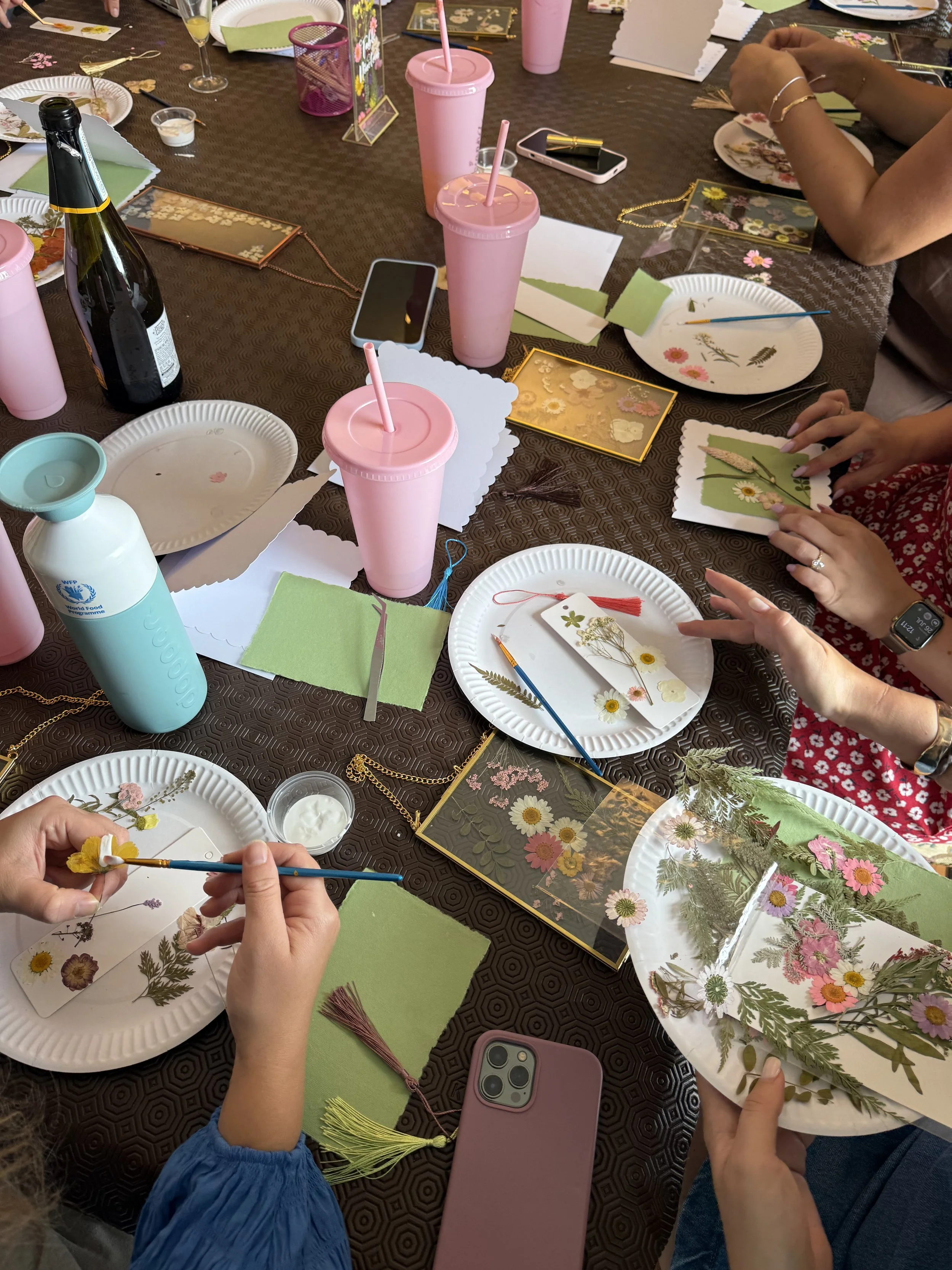 People decorating handmade floral greeting cards at a craft party, using pressed flowers and paint on paper plates, with drinks and crafting supplies on the table.