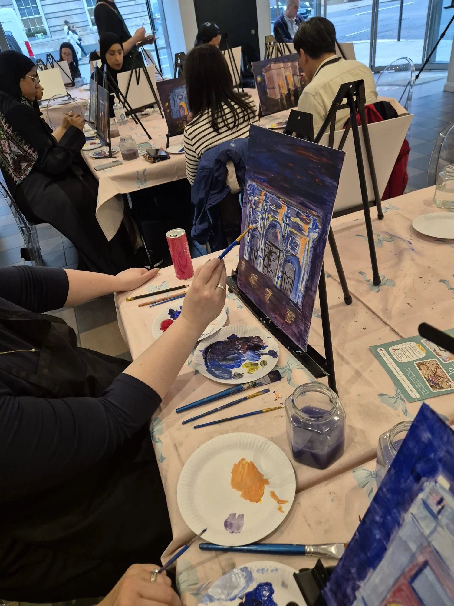 People participating in a group painting class indoors, with canvases on easels, art supplies, and a view of the street outside through large windows.