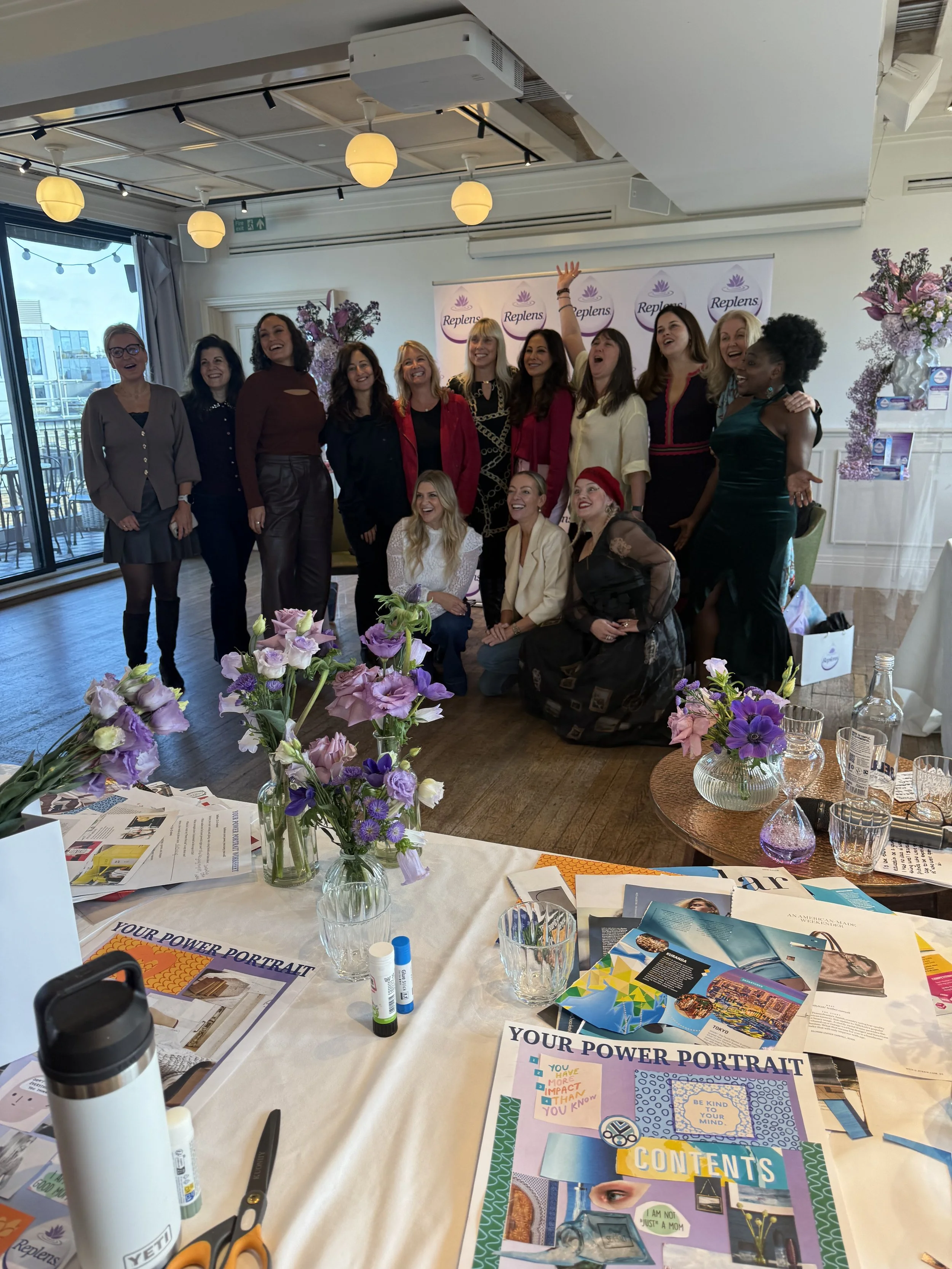 A group of women posing for a photo at a women's event, with purple flowers and event banners in the background, and a table with flowers, brochures, and craft supplies in the foreground.