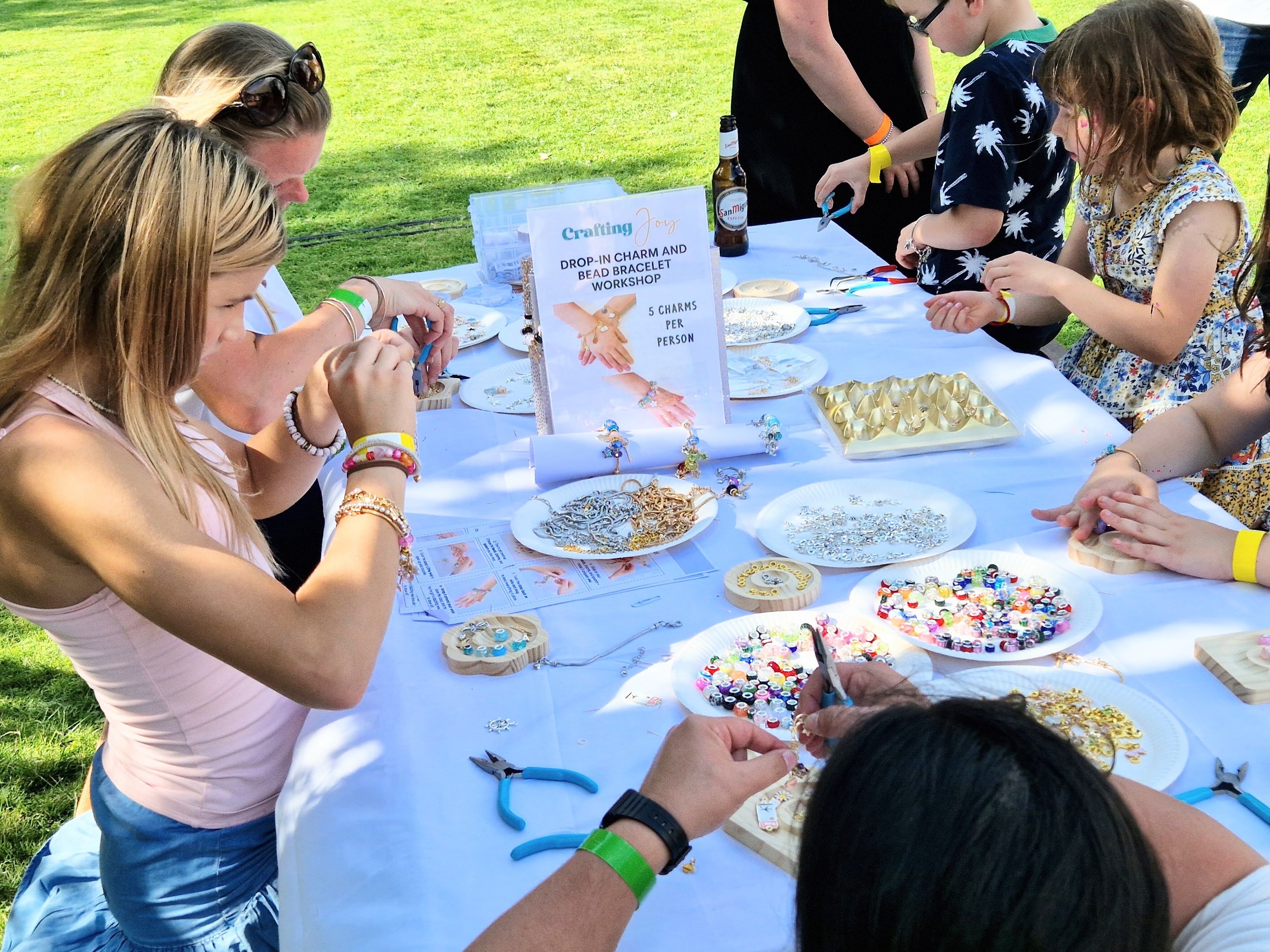 Children participating in a bead bracelet-making workshop at an outdoor craft fair, with supplies like beads, charms, and tools on a table.