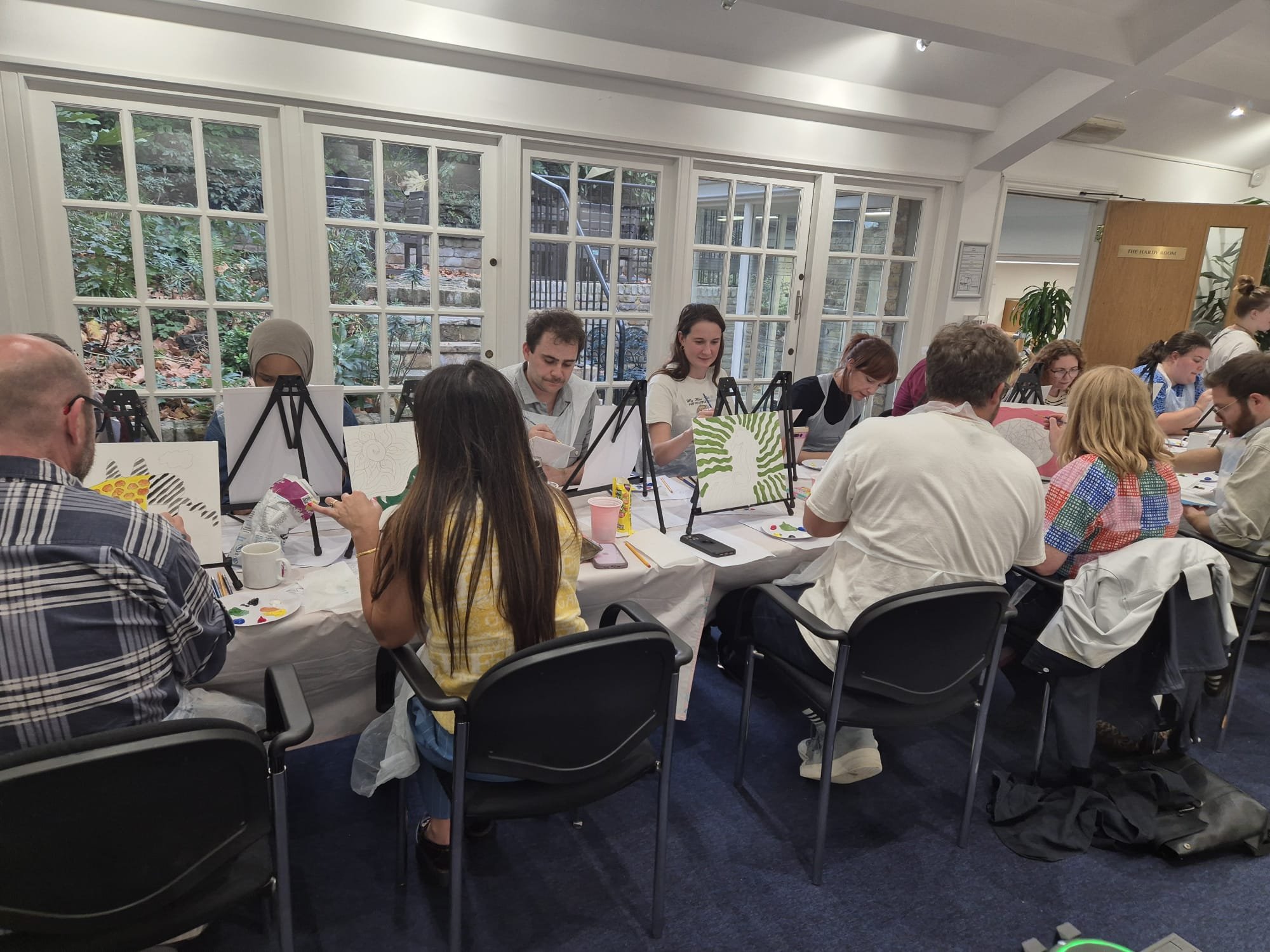 Group of people sitting at a long table painting canvases with abstract and patterned designs during an art class in a bright room with large windows.