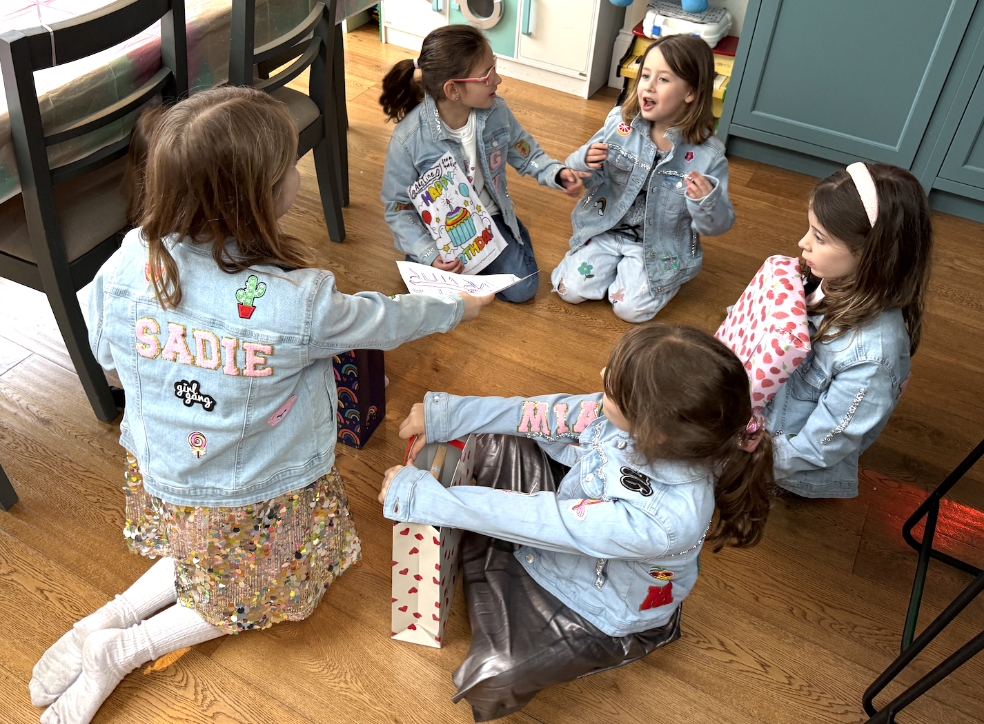 Six young girls having a birthday celebration indoors. They are sitting or kneeling on a wooden floor, wearing denim jackets with colorful patches and embroidery, and exchanging gifts. One girl is holding a birthday card, while another girl is openin