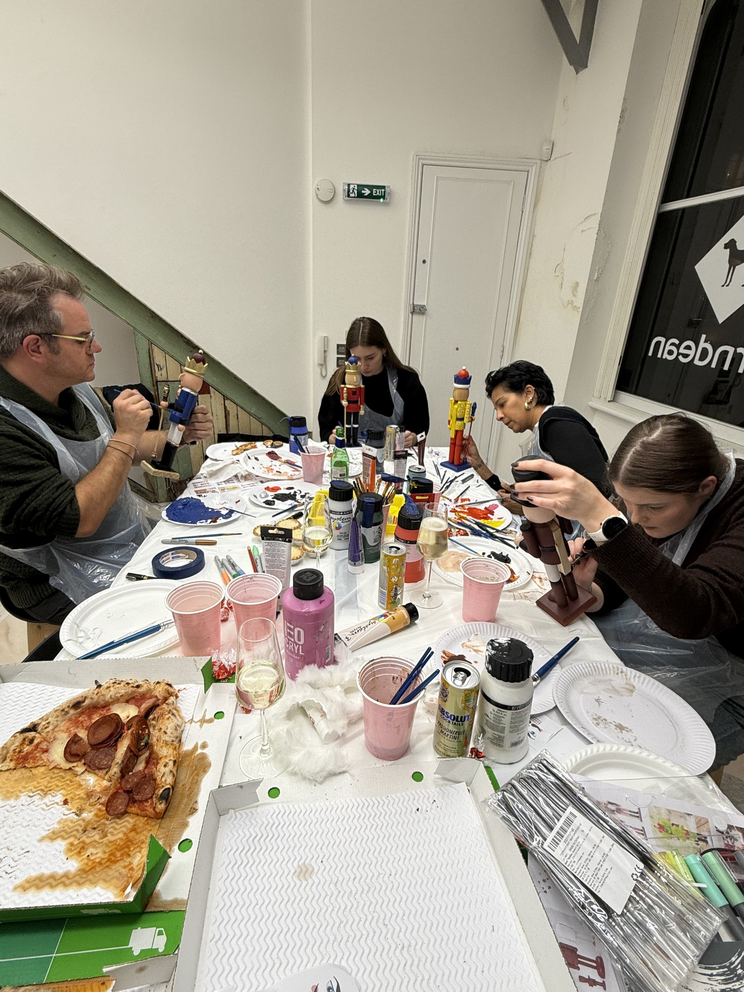 People painting nutcracker figurines at a table covered with art supplies, pizza, and drinks, with a white wall and door in the background.