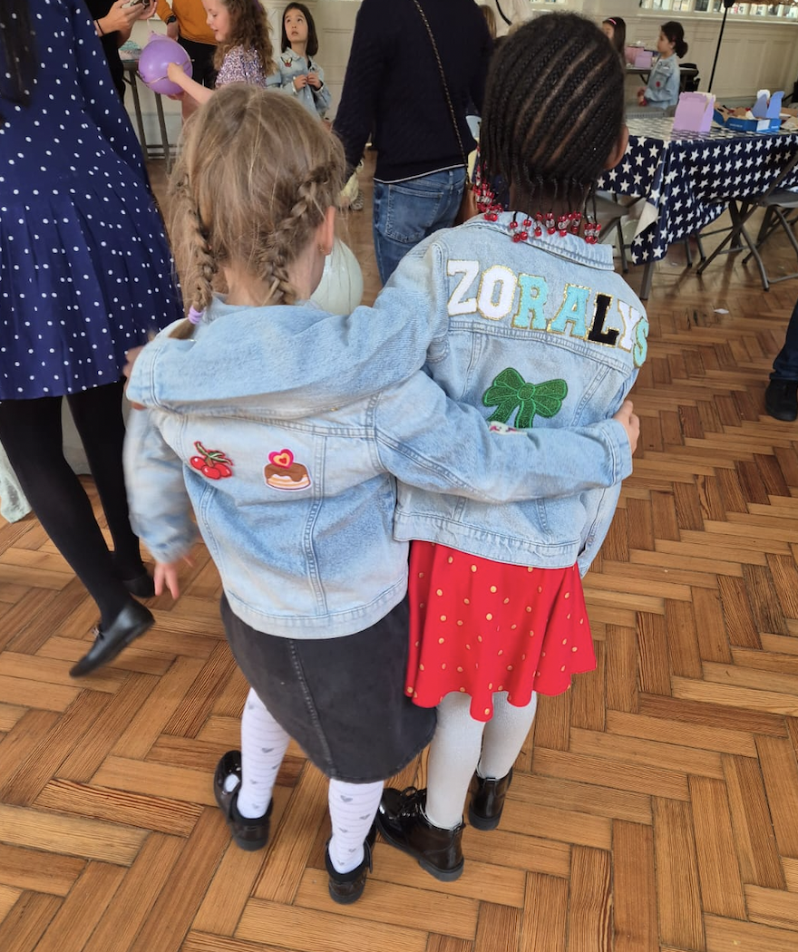 Two young girls hugging at a party or gathering. One girl has blonde braids, a denim jacket with patches, a black skirt, and white socks. The other girl has braided hair, a denim jacket with the name 'Zorys' and a green bow patch, a red polka dot dre