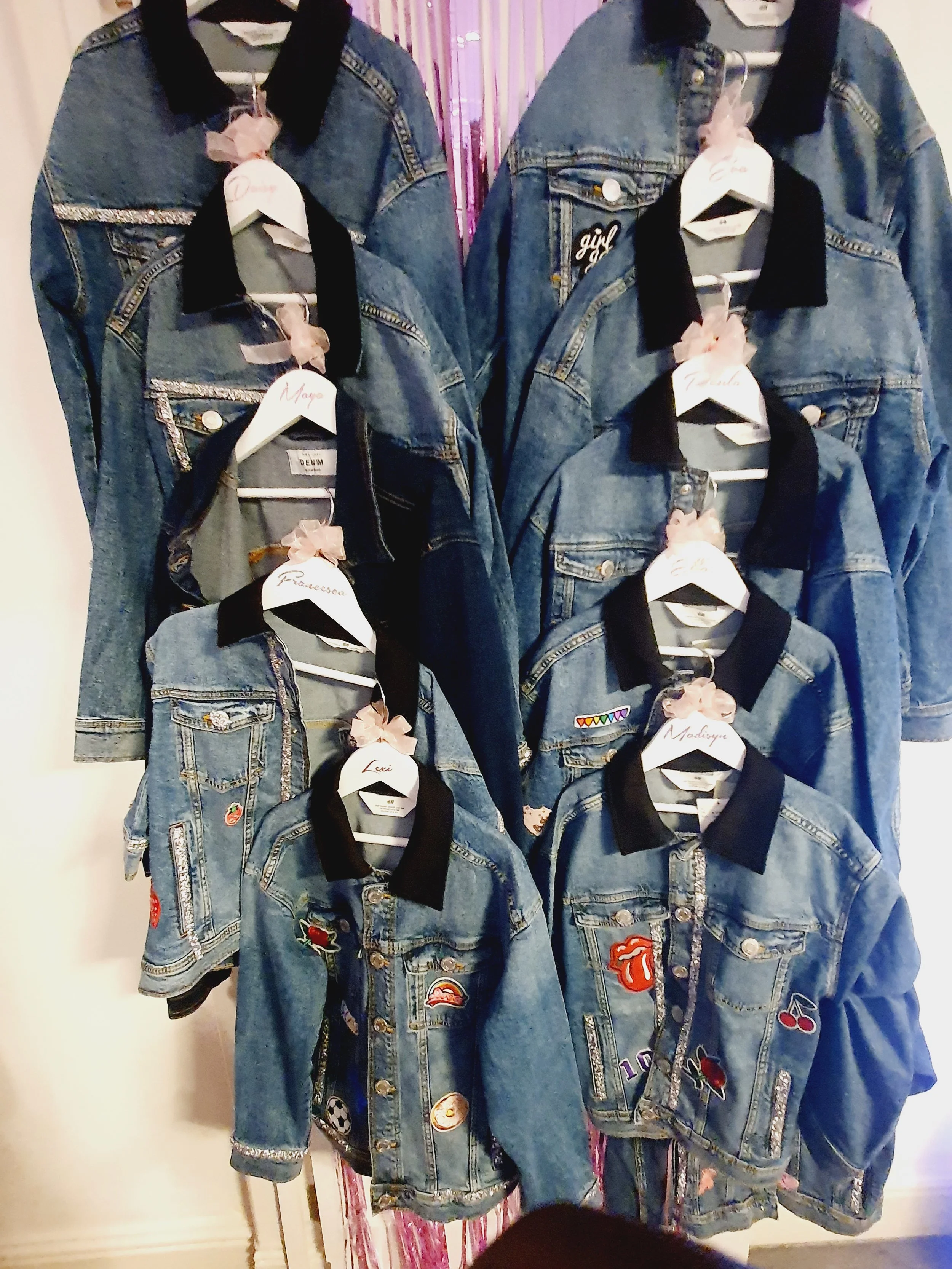 A display of eight denim jackets with various patches and embellishments, hung on white hangers with pink bows, arranged in two columns against a background of pink and purple vertical stripes.