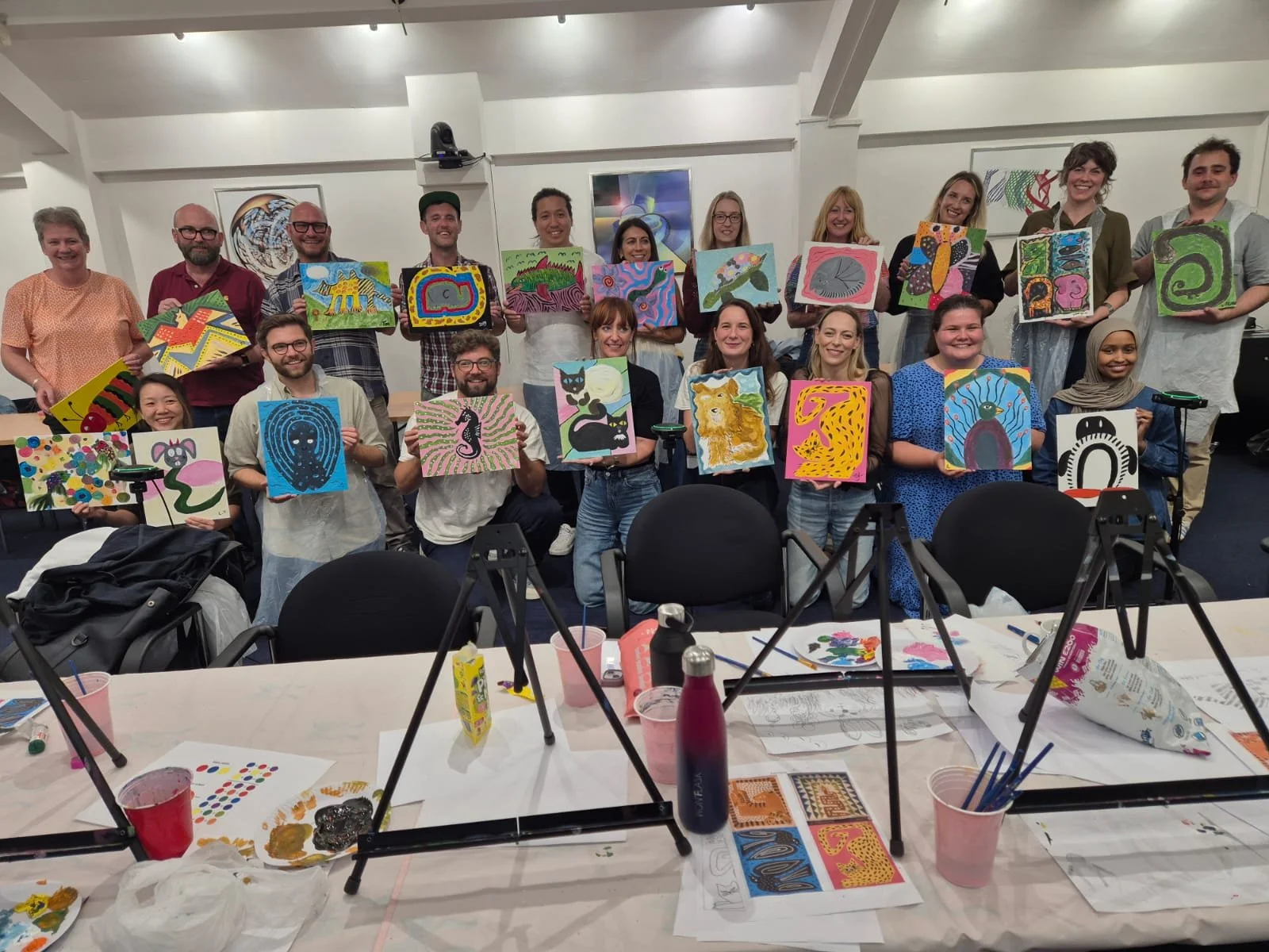 Group of people holding colorful abstract paintings in an art class or workshop, with art supplies and paintings on a table in the foreground.