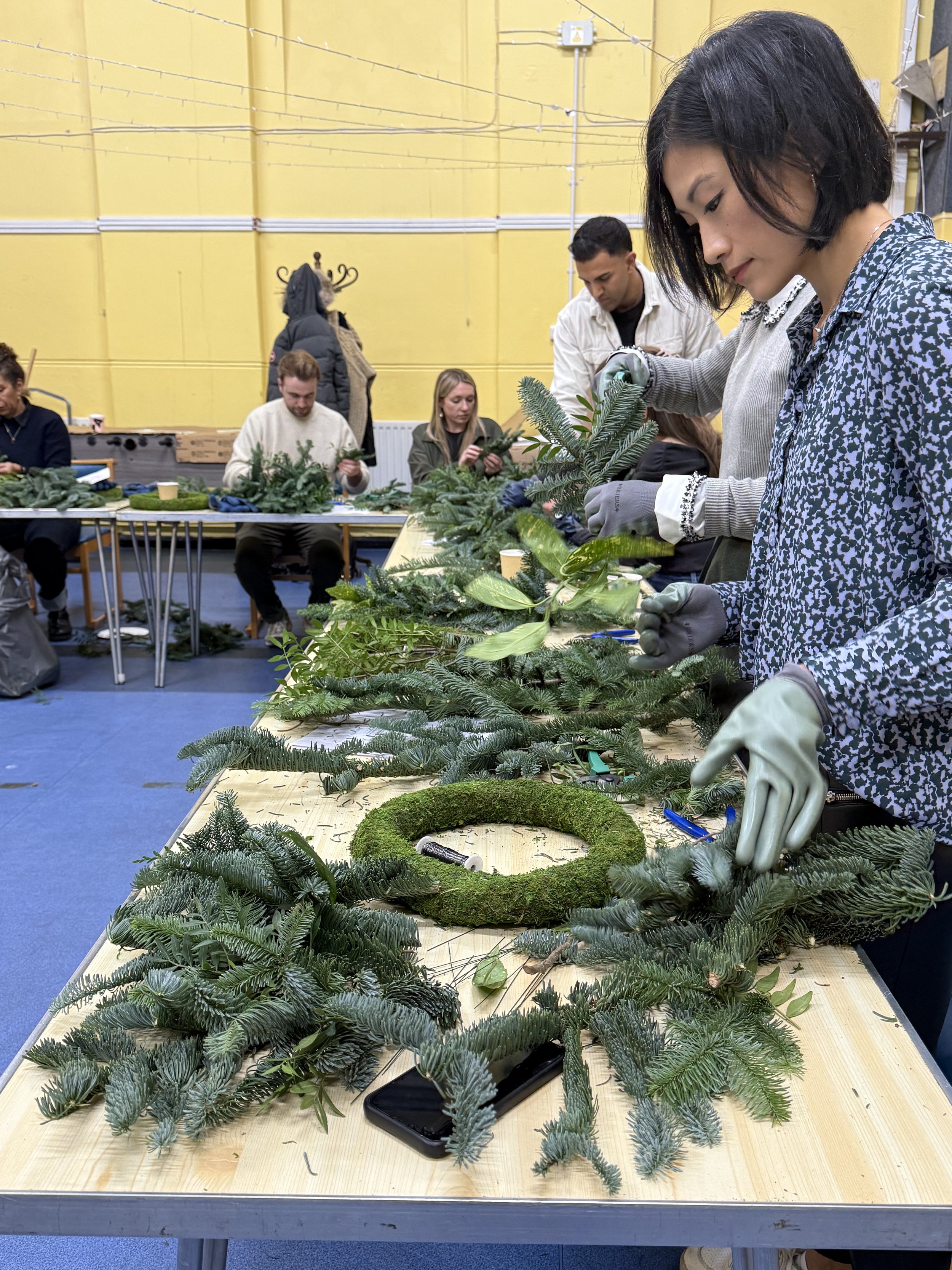 People crafting holiday wreaths and arrangements using pine branches and greenery in a workshop with yellow walls.
