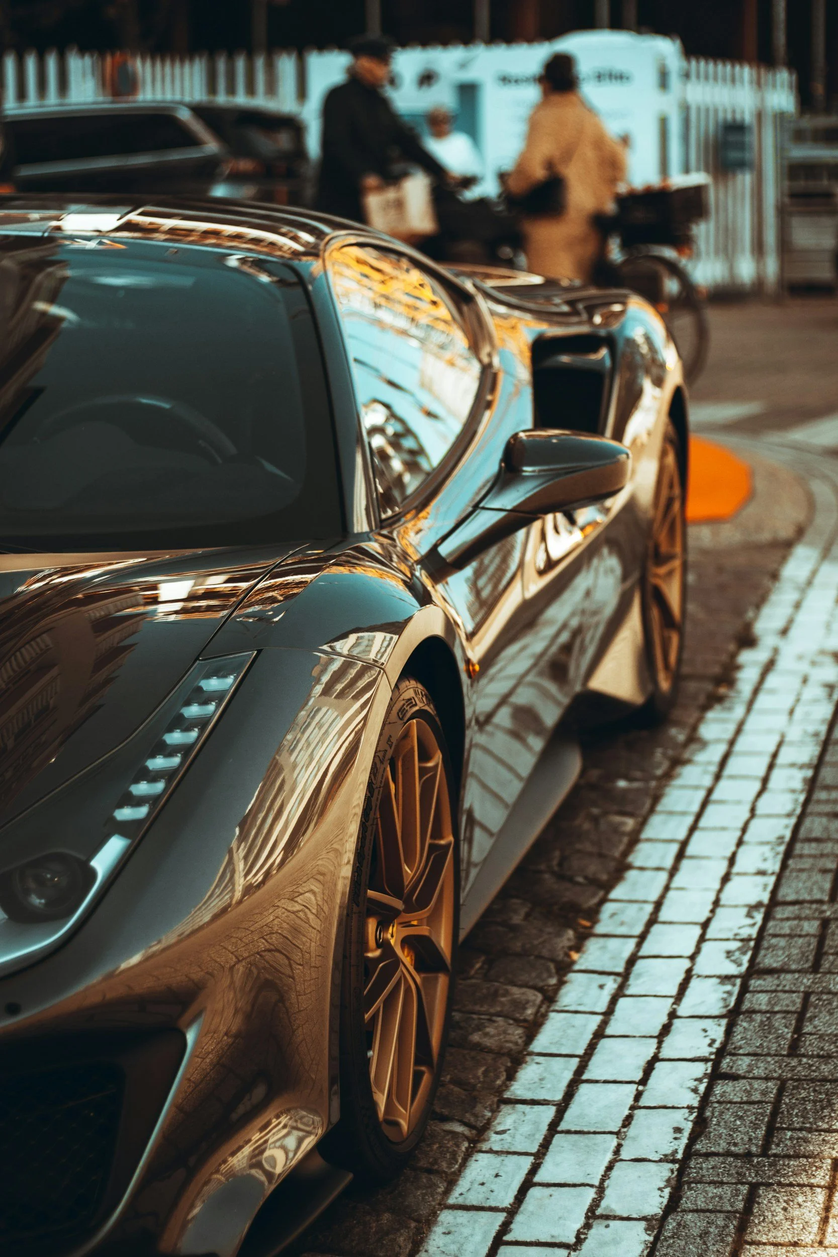 Close-up of a parked gray sports car on a city street, with reflections of buildings on its shiny surface, and people with delivery bikes in the background.
