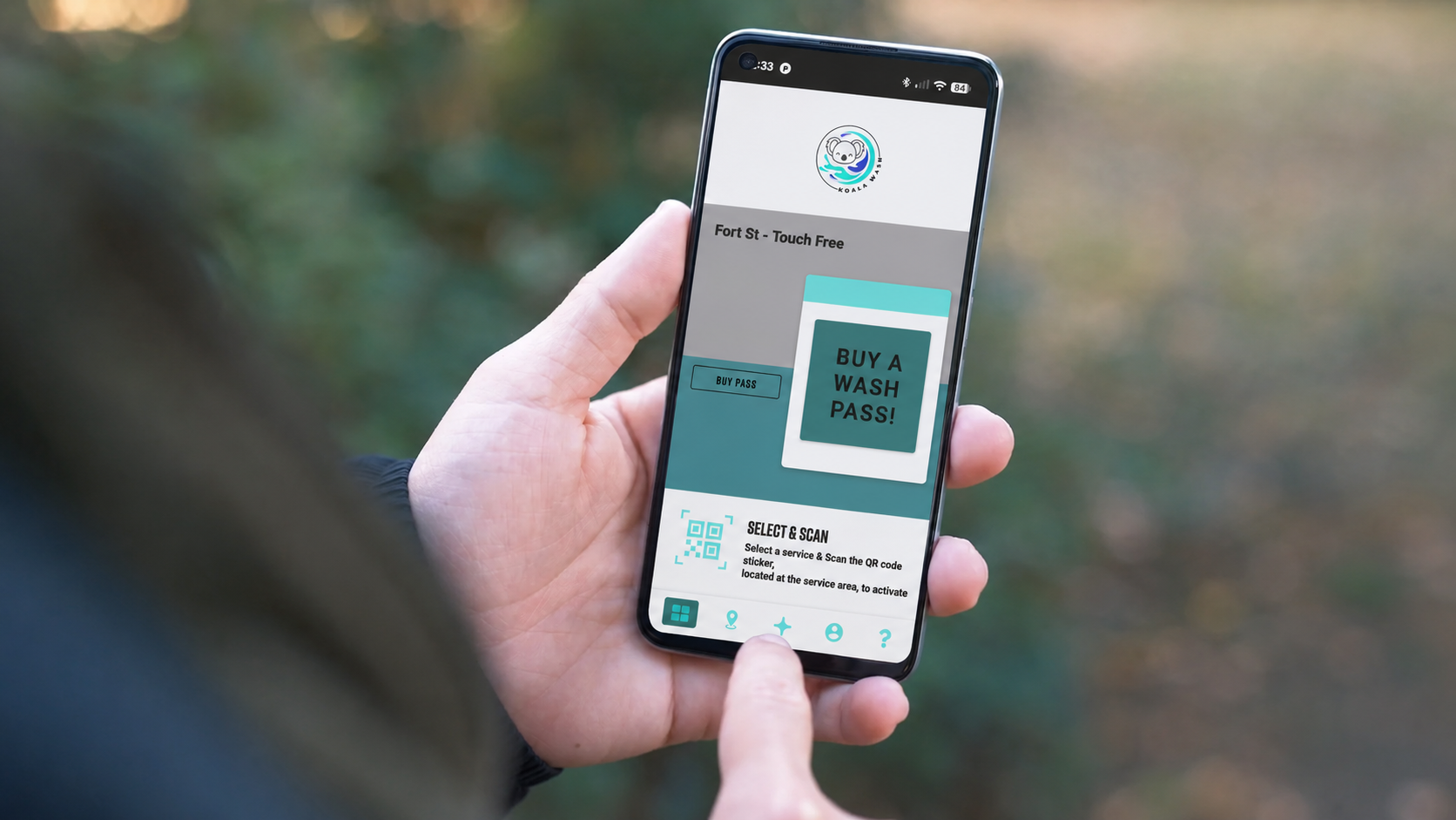 Person holding a smartphone displaying a Koala Way wash PASS app, with options to buy pass and scan QR code, outdoors with blurred greenery background.