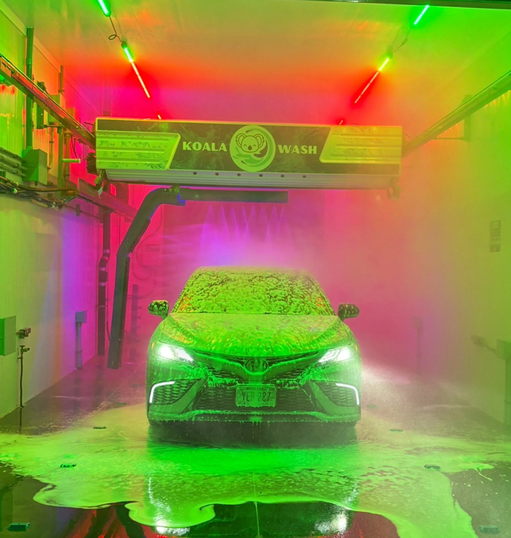 A car in a car wash with colorful lights and water spray, with a sign reading 'Koala Wash' and a koala logo.