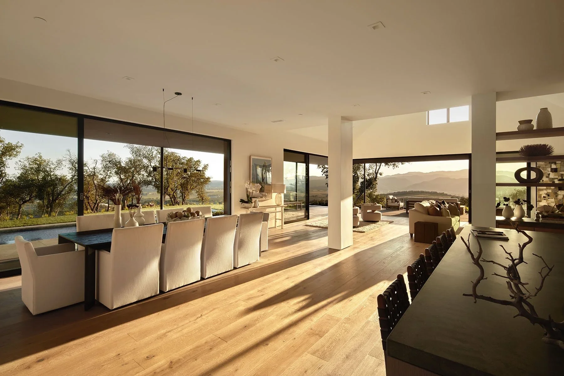 A modern, open-concept living and dining area with large floor-to-ceiling windows showing a scenic view of trees and mountains at sunset. The space features a long dining table with white chairs, a sitting area with sofas, decorative vases and artwor