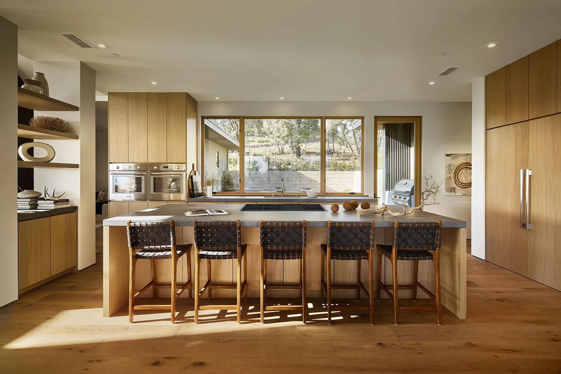 Modern kitchen with wooden cabinets, large window, and island with seating for five, decorated with branch and pottery, bright natural light.