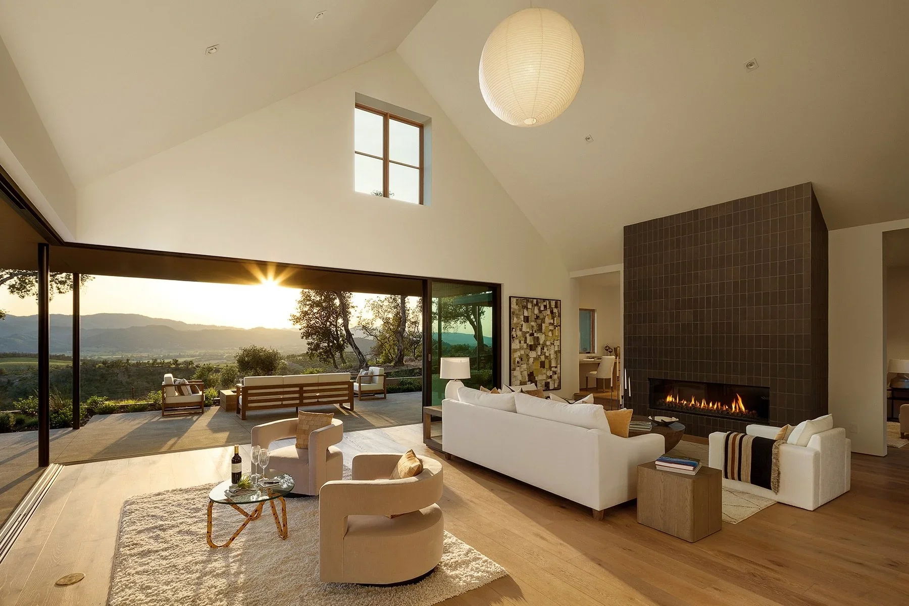 Spacious living room with large glass doors opening to outdoor patio and scenic landscape. Interior features white sofas, armchairs, a fireplace, and modern decor. Sunset visible through the glass doors.