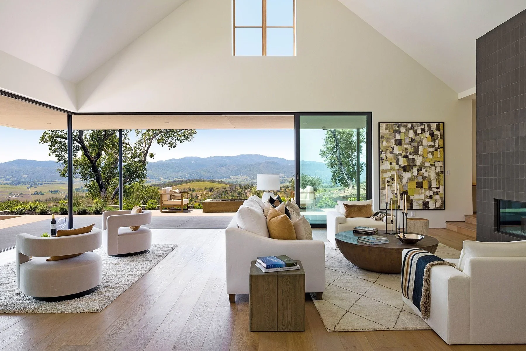 Modern living room with large glass sliding doors opening to scenic outdoor view of mountains and trees, white sofas with beige pillows, a circular wooden coffee table, an abstract wall art, and a textured grey fireplace.