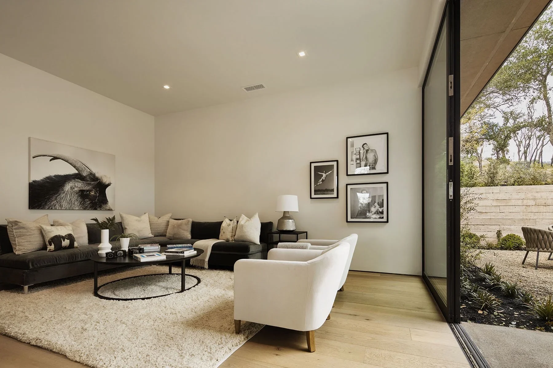 Living room with black sectional sofa, white armchair, beige rug, and wall art including a goat photograph, with large sliding glass door opening to an outdoor patio and garden.