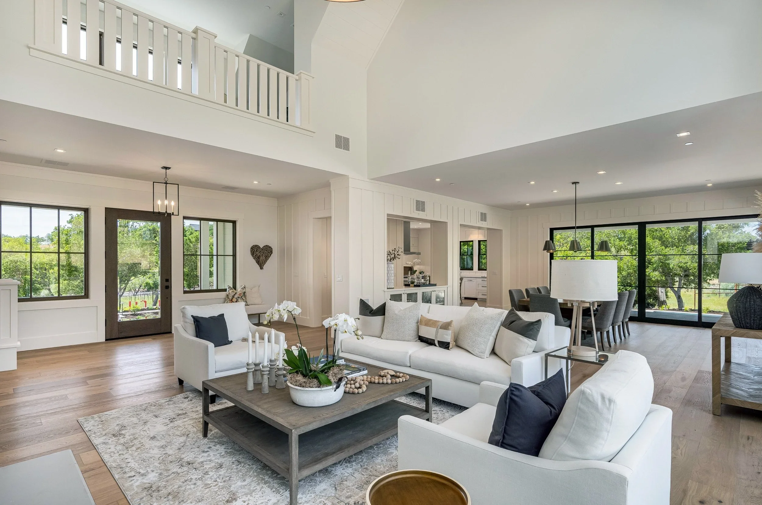 Open concept living room with white sofas, wooden coffee table, and large windows showing greenery outside, in a modern bright house.
