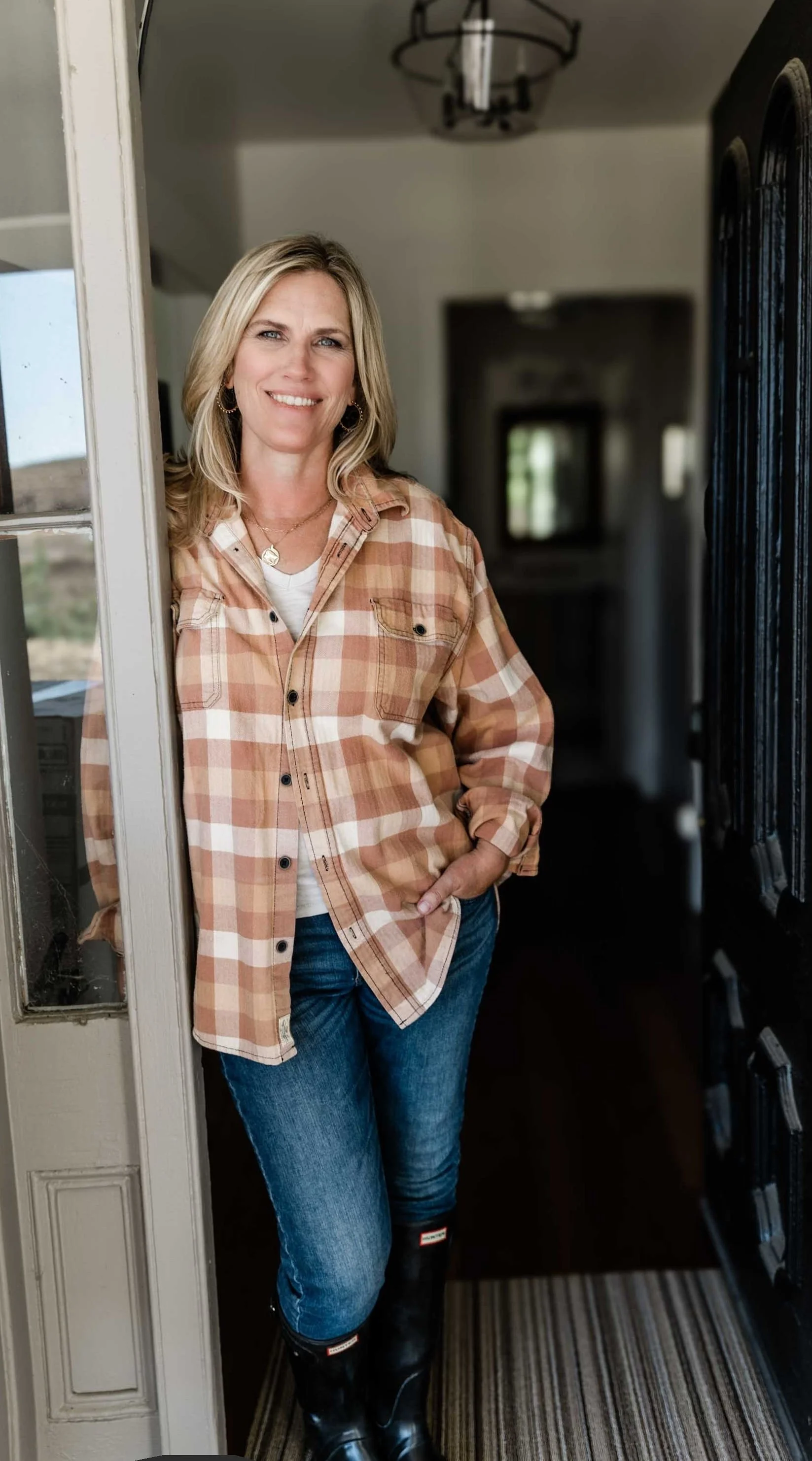Woman with blonde hair, wearing a plaid shirt, jeans, and boots, standing in a doorway inside a home, smiling at the camera.
