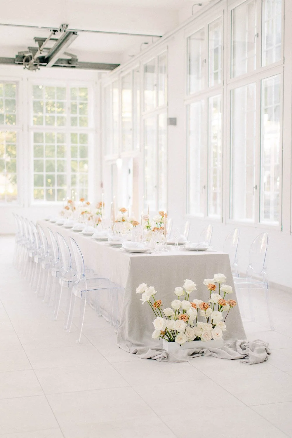 Simple wedding table setup on a long indoor table with neutral linen tablecloth, taper candles, glassware, ghost chairs and soft rose florals by de Waay
