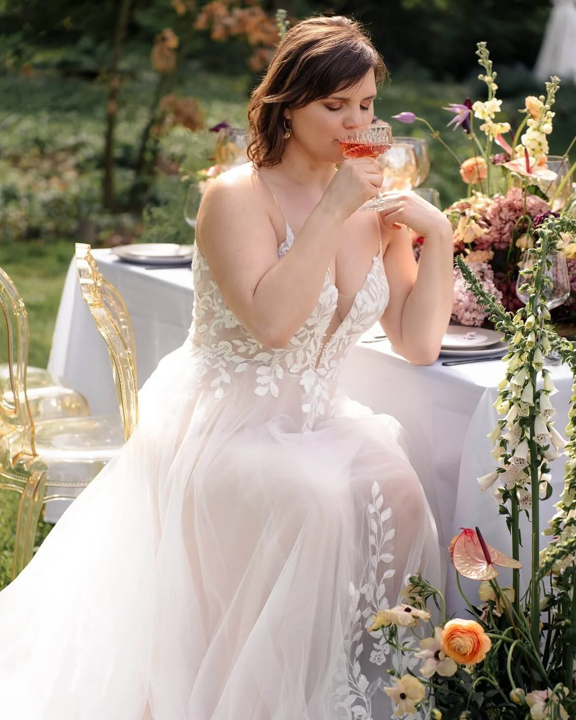 Braut sitzt an frühlingshaft gedeckter Hochzeitstafel mit zarter Floristik, Ghost Chairs von de Waay und warmem Licht – Tischdeko Hochzeit Frühling in natürlicher, moderner Atmosphäre