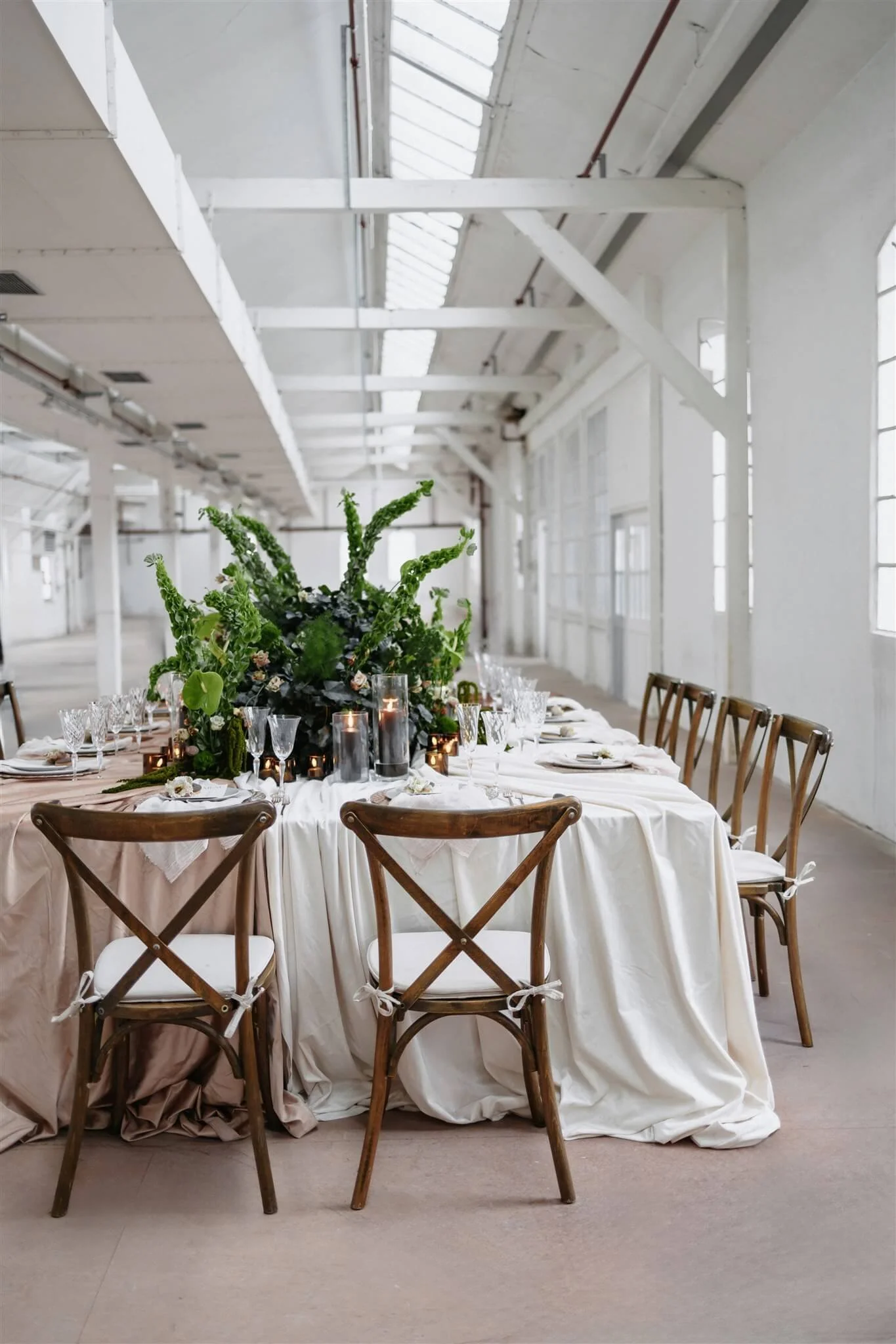 Elegante Hochzeitstafel von de Waay mit warmen Neutraltönen, grüner leichter Floristik und Crossback Stühlen in einer lichtdurchfluteten Industriehalle – Hochzeitsfarben 2026
