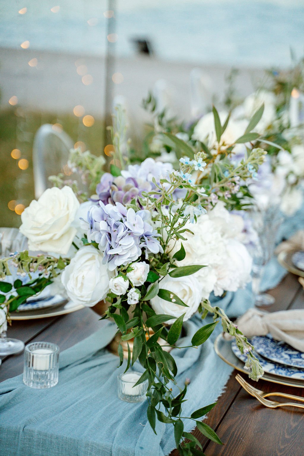 Tischdeko Hochzeit Blau: Blumengesteck aus Eukalyptus, weißen Rosen und pastellblauen Hortensien auf Leinentuch in Dusty Blue – edel und natürlich zugleich de Waay