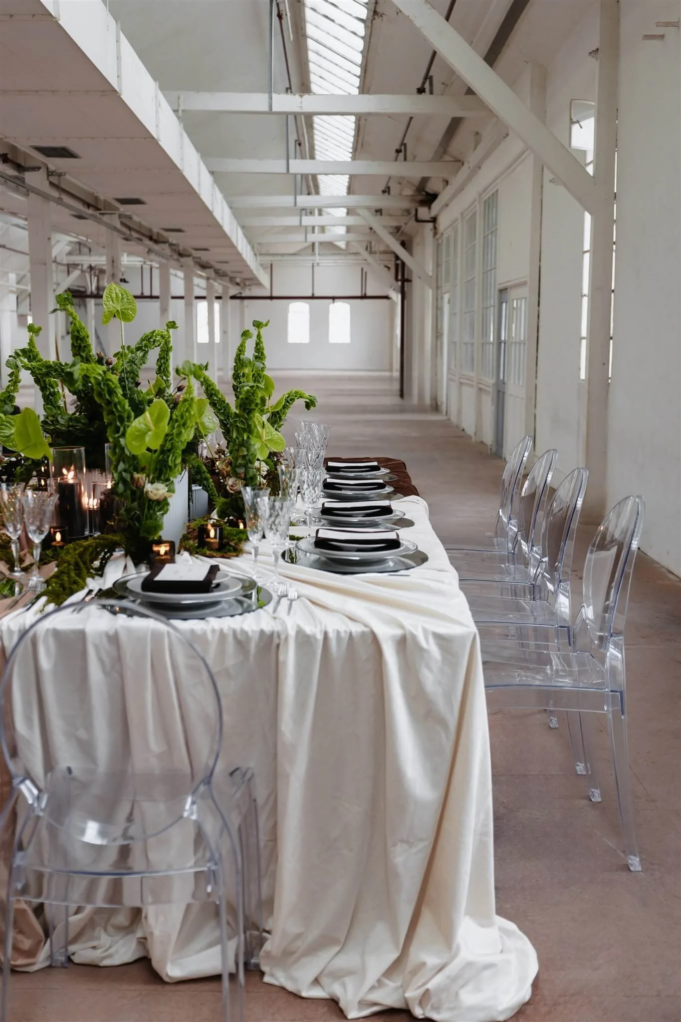 Hochzeitsfarben 2026: elegante Hochzeitstafel mit Ghost Chairs, silbernen Platztellern, Kristallgläsern und silbernem Besteck von de Waay mit Tischdecken in warmen Neutrals in einer hellen Industriehalle
