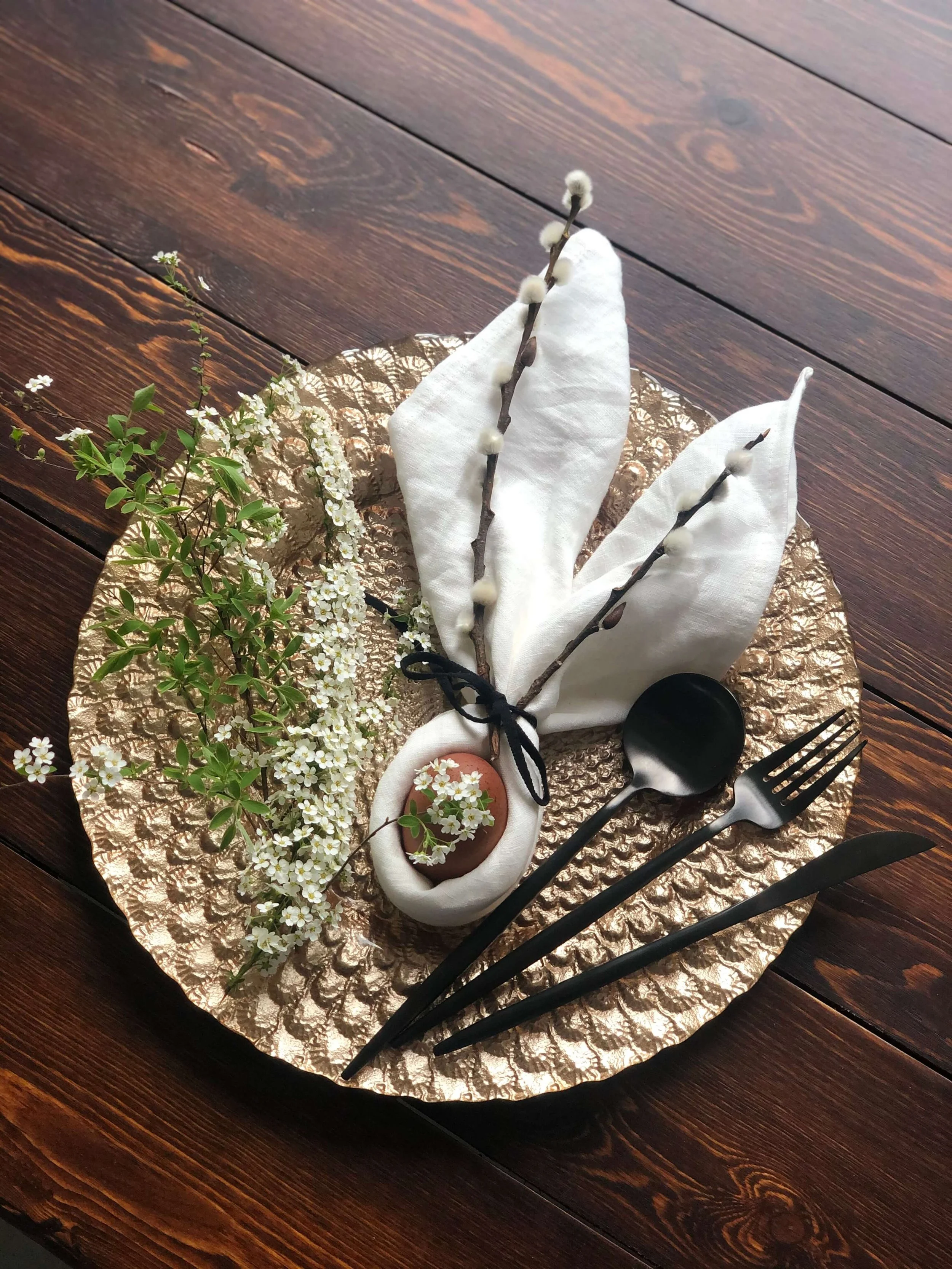 Ostergedeck mit goldenem Platzteller, Leinenserviette, Ei, Zweig und schwarzem Besteck auf Holztisch