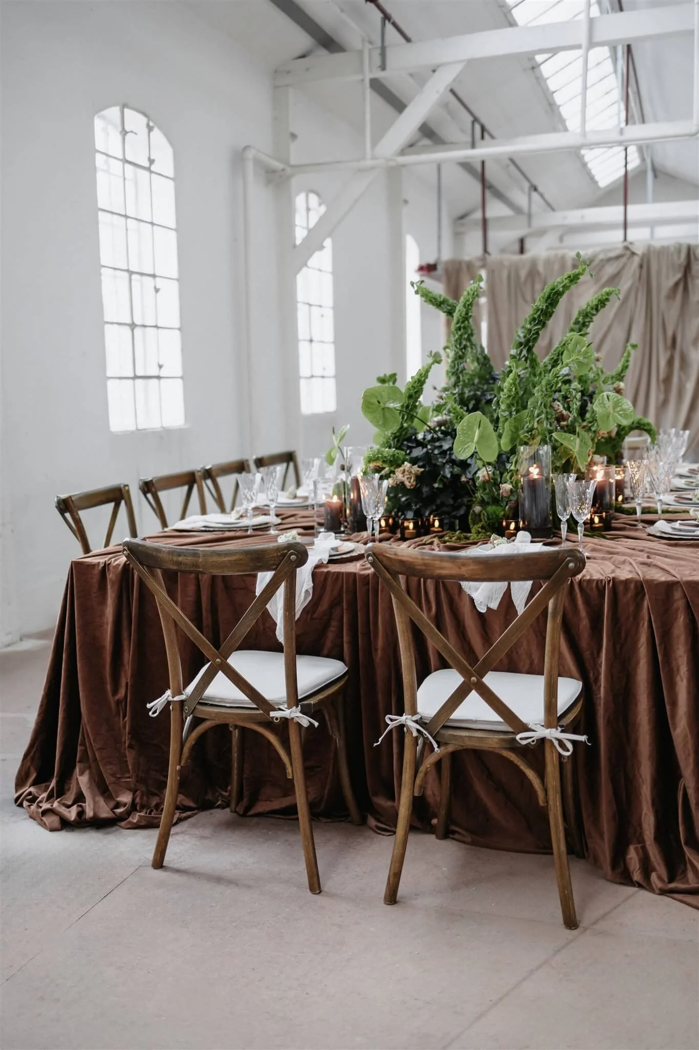 Hochzeitstafel mit Crossback Stühlen in warmen Brauntönen mit grüner Floristik, Kupferbesteck und Cappucchinofarbigen Platztellern von de Waayund Samttischdecke – Hochzeitsfarben 2026 in einer modernen Industriehalle