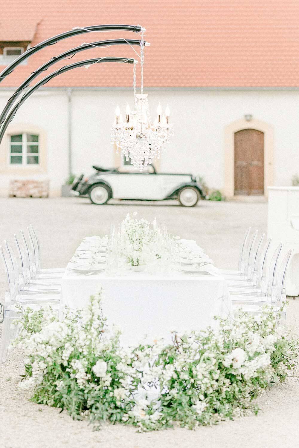 All White Cloud Dancer Tischdeko zur Hochzeit mit Ghost Chairs, weißer Tafel, Kronleuchter und floraler Installation von de Waay – Hochzeitsfarben 2026
