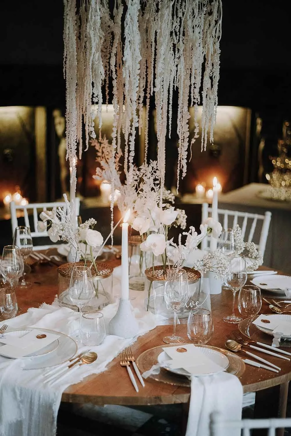 Weiße Tischdeko zur Hochzeit mit Holztisch, weißen Tiffany Stühlen, warmem Kerzenlicht, Goldbesteck und natürlicher All-White-Floristik von de Waay