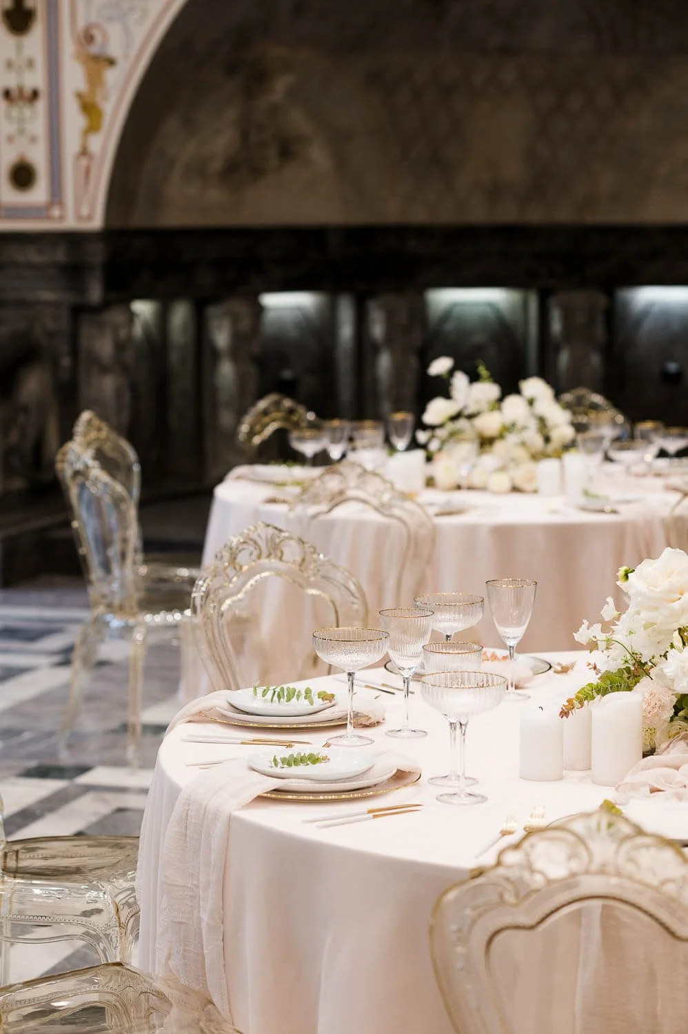 Simple elegant round wedding table decor with ivory tablecloth, low floral centerpieces, candles, gold accents and transparent chairs by de Waay