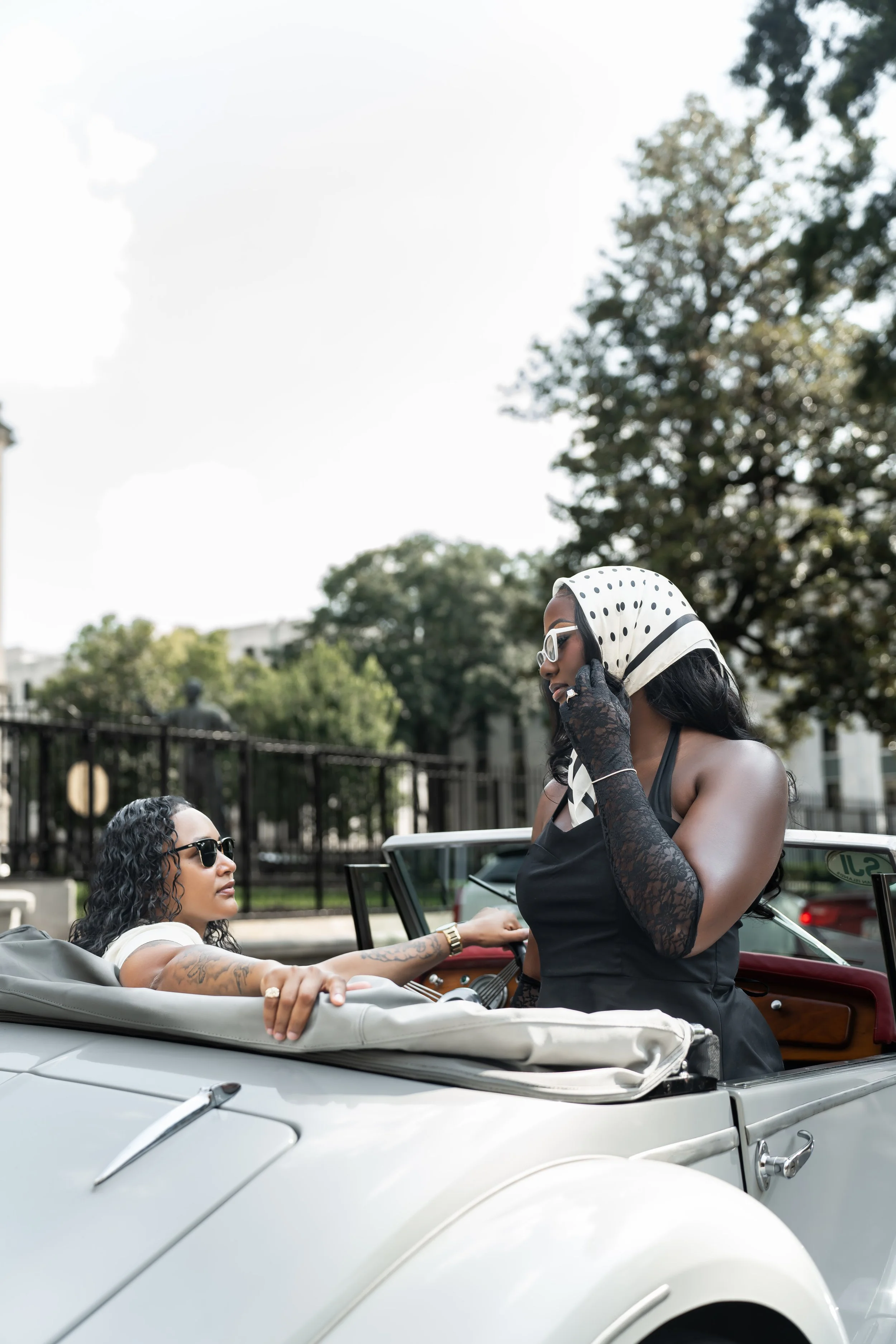 Two women in sunglasses are in a vintage convertible car, one standing and talking on a cellphone, the other sitting and looking at her.