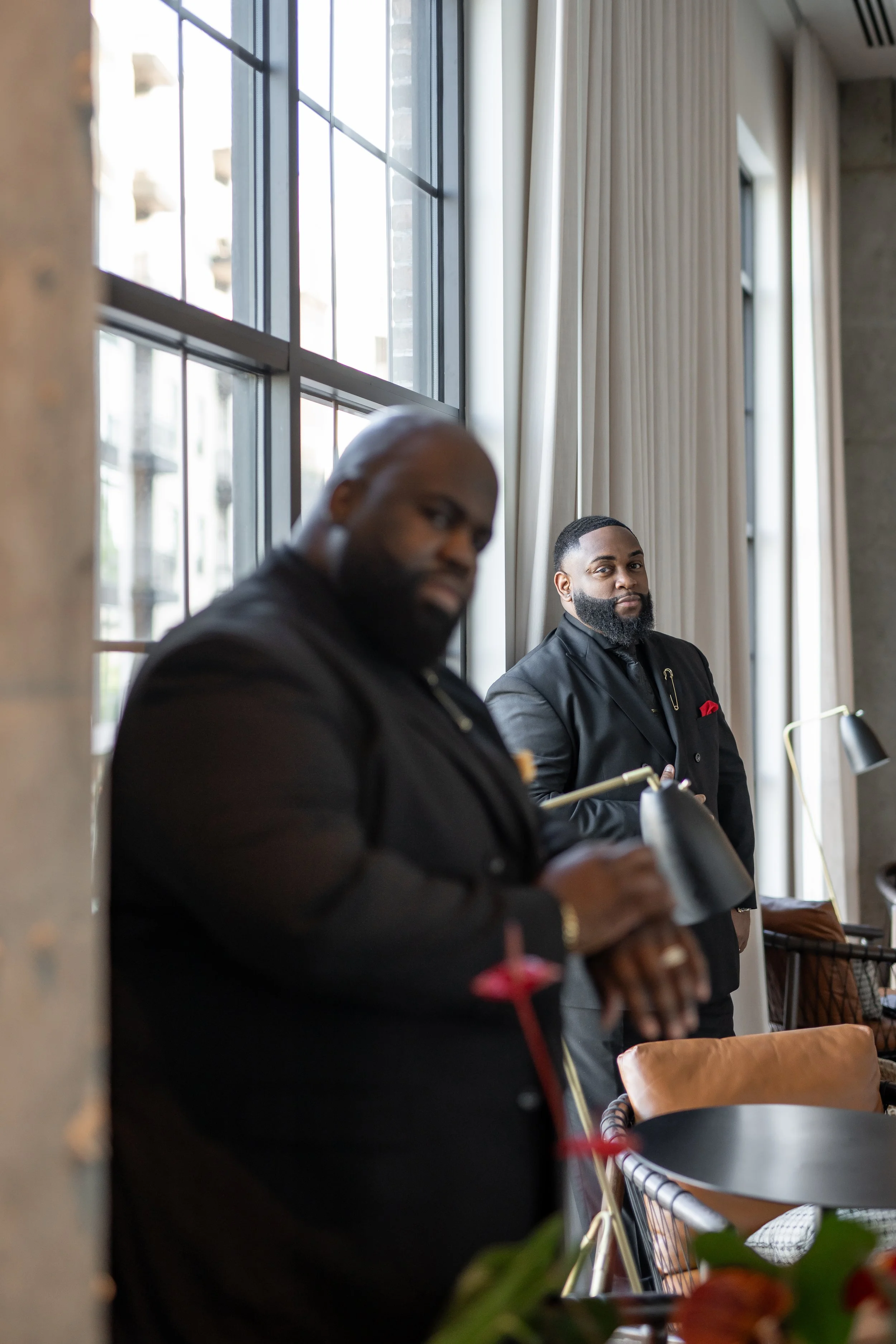 Two well-dressed men with beards standing next to a large window with white curtains, in a well-lit room with a modern industrial style interior.