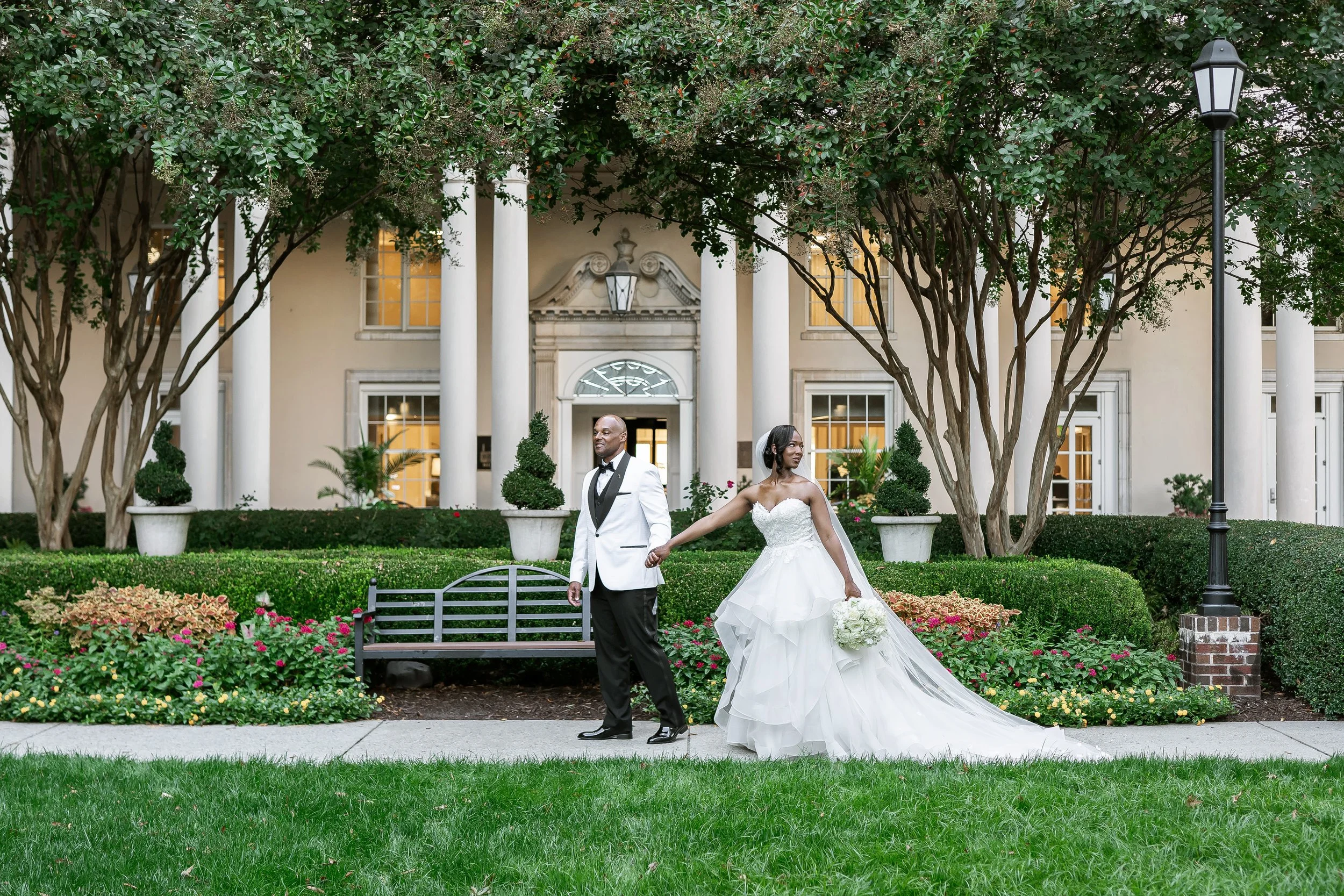 A bride and groom walking hand in hand in front of a large house with white columns, surrounded by greenery and flowers.