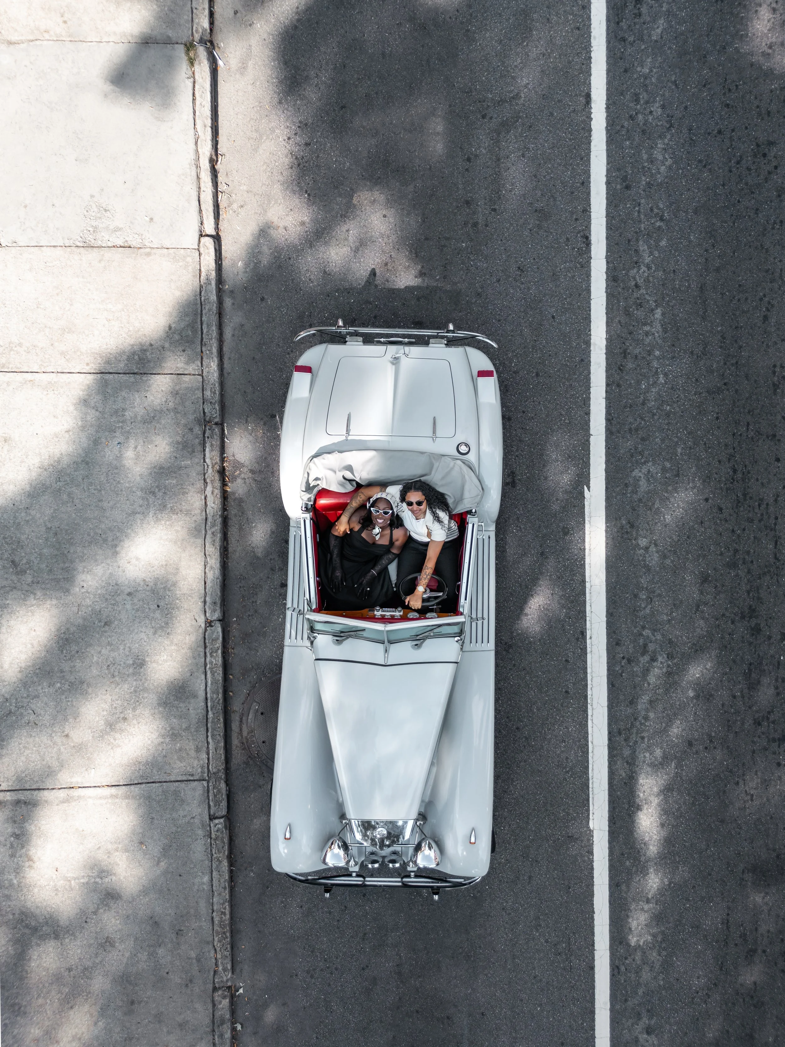 An aerial view of two women sitting in a vintage convertible car parked on dark asphalt, under the shade of trees.