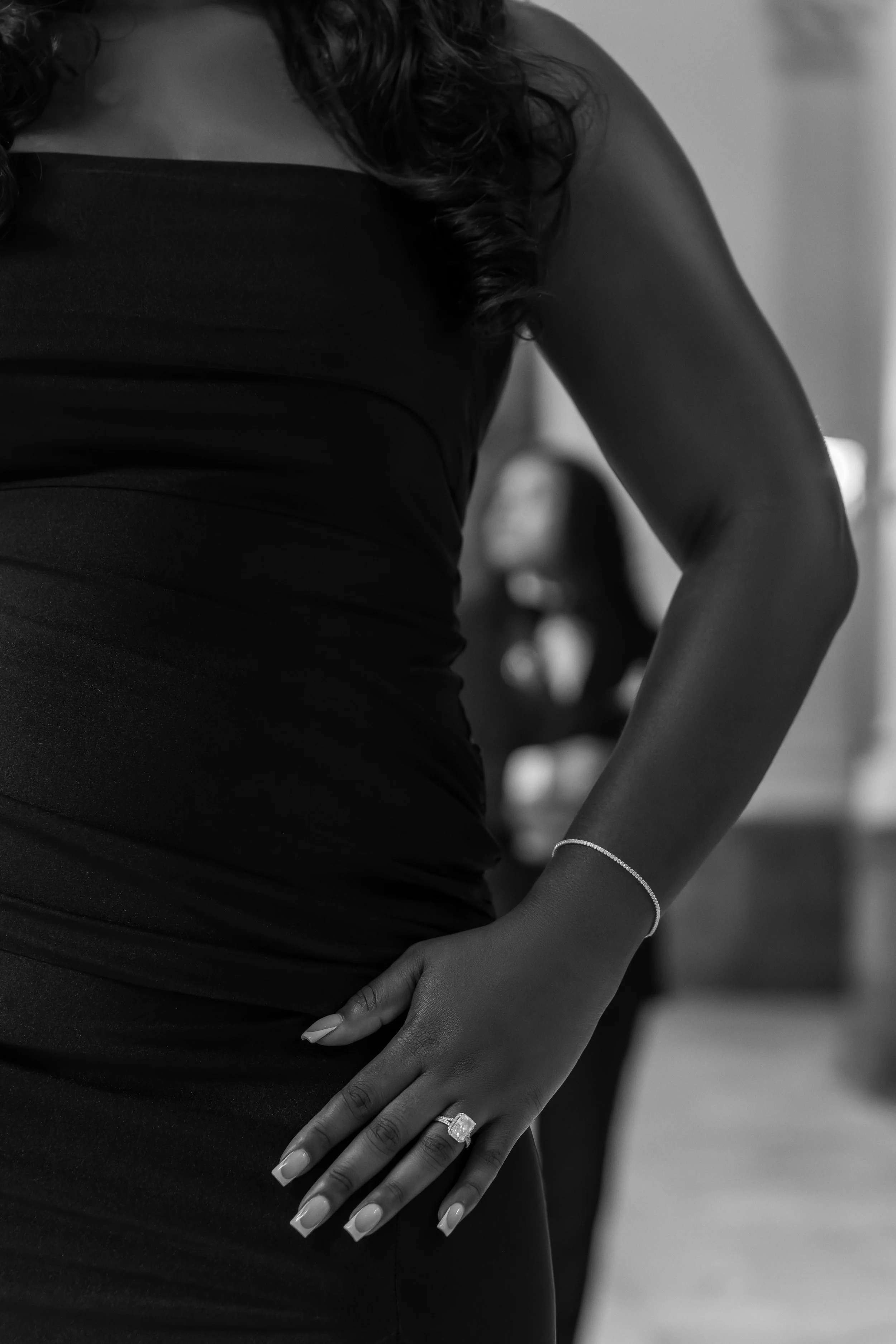 Close-up of a woman in a black dress, showing her left hand on her hip with a large engagement ring and a bracelet on her wrist.