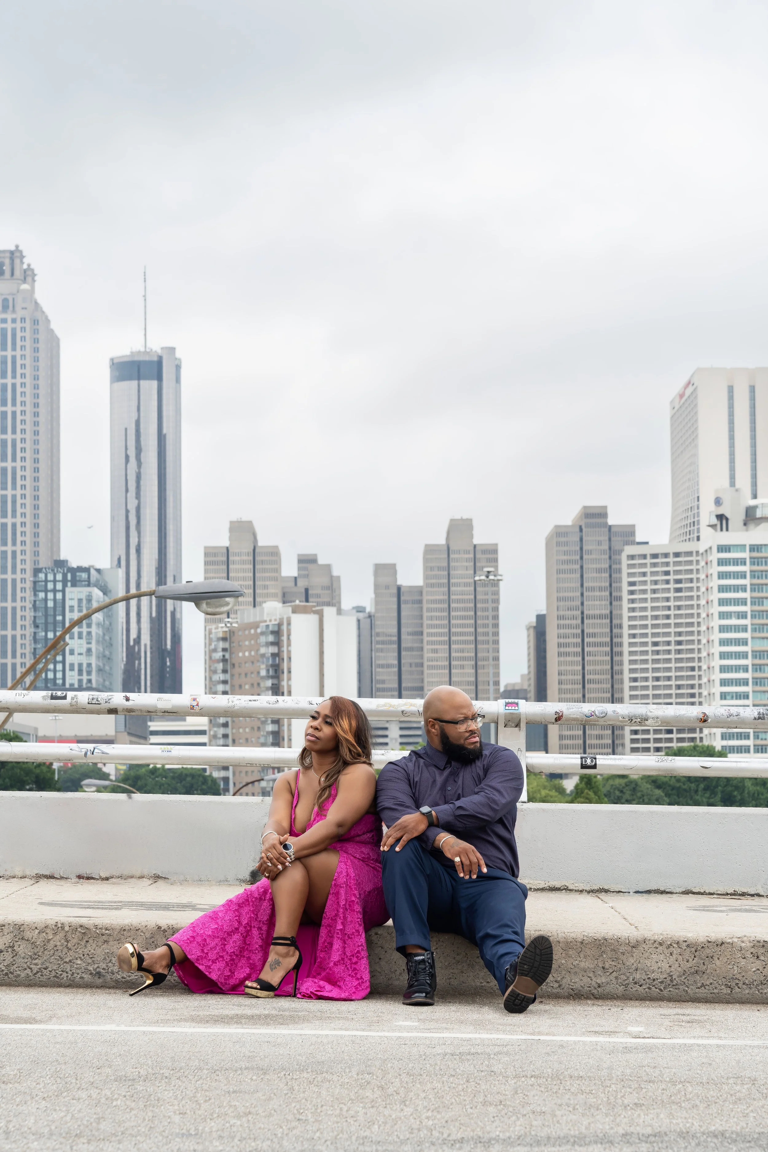 A man and woman sitting on the curb of a city street, leaning against each other, with a city skyline in the background.