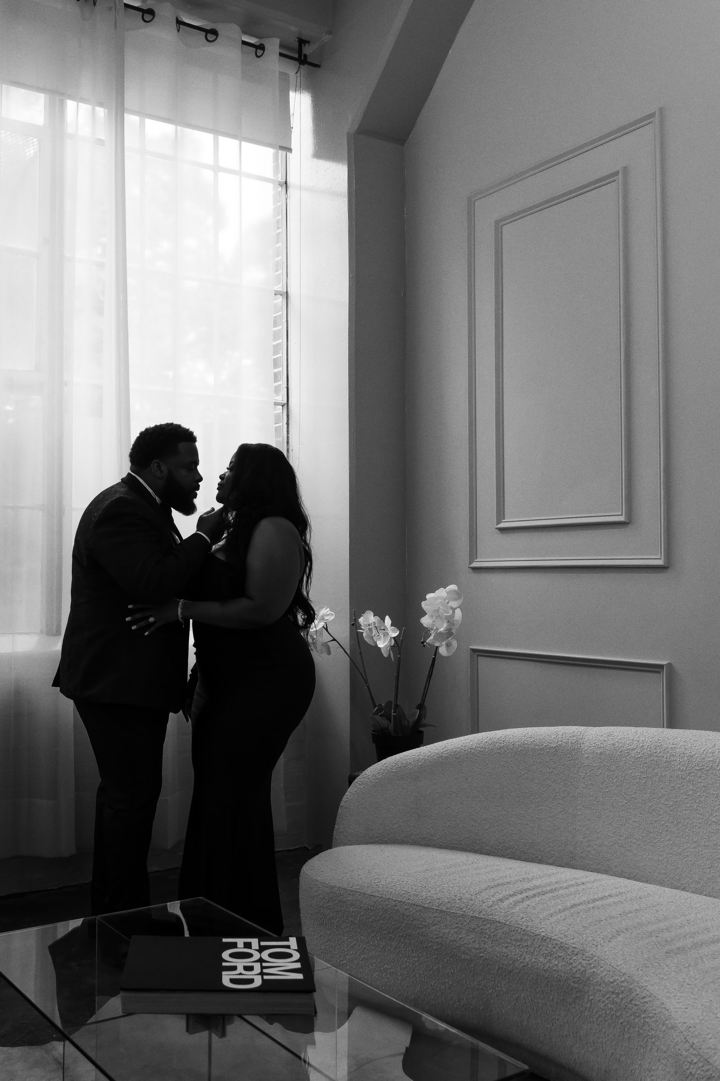 A black and white photo of a couple standing close together near a window with sheer curtains, inside a room with a white couch, a glass table with a book on it, and a potted plant with white flowers.