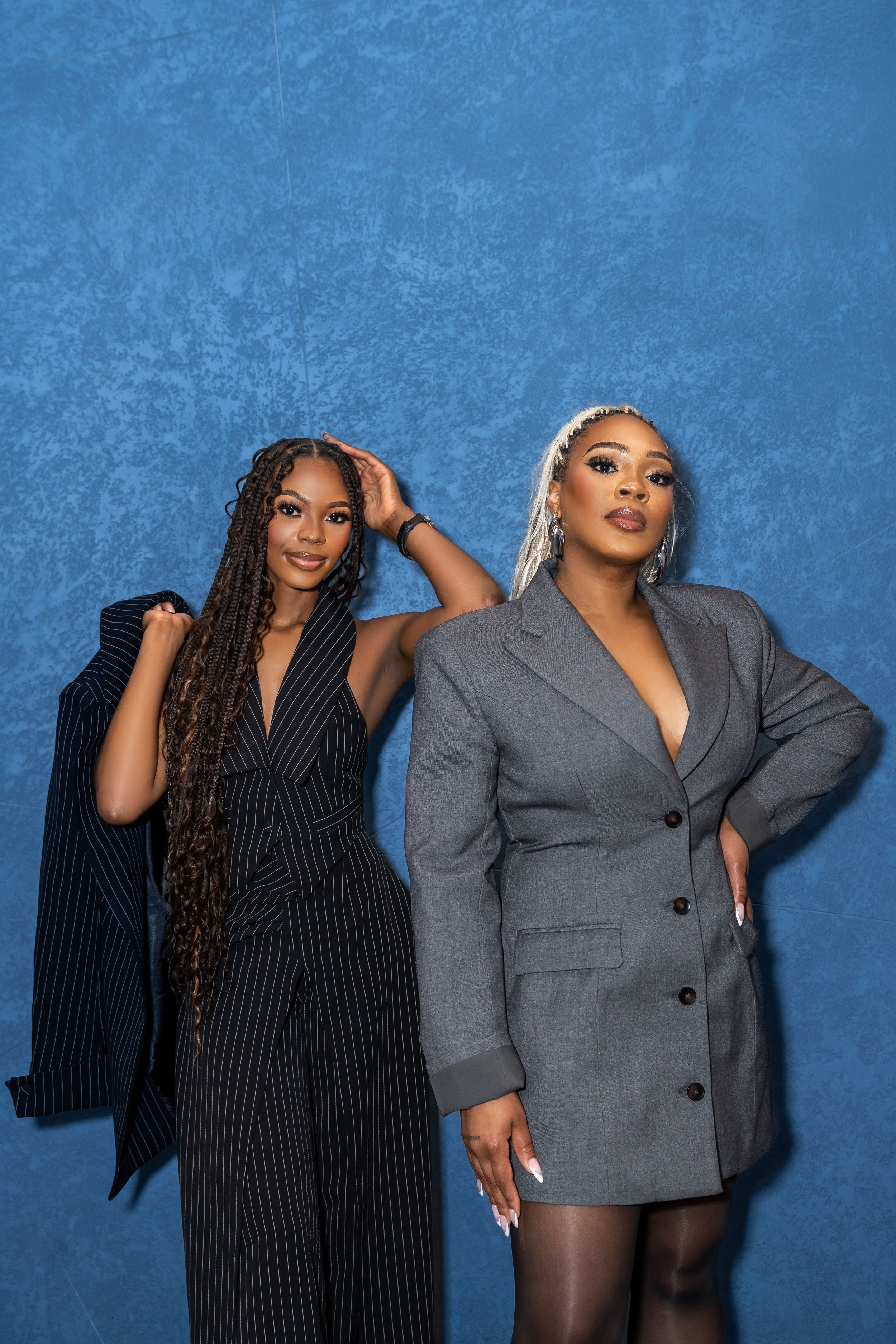 Two women posing in front of a blue textured wall, one wearing a black pinstripe suit with a blazer over her shoulder, and the other in a gray suit jacket with black stockings.