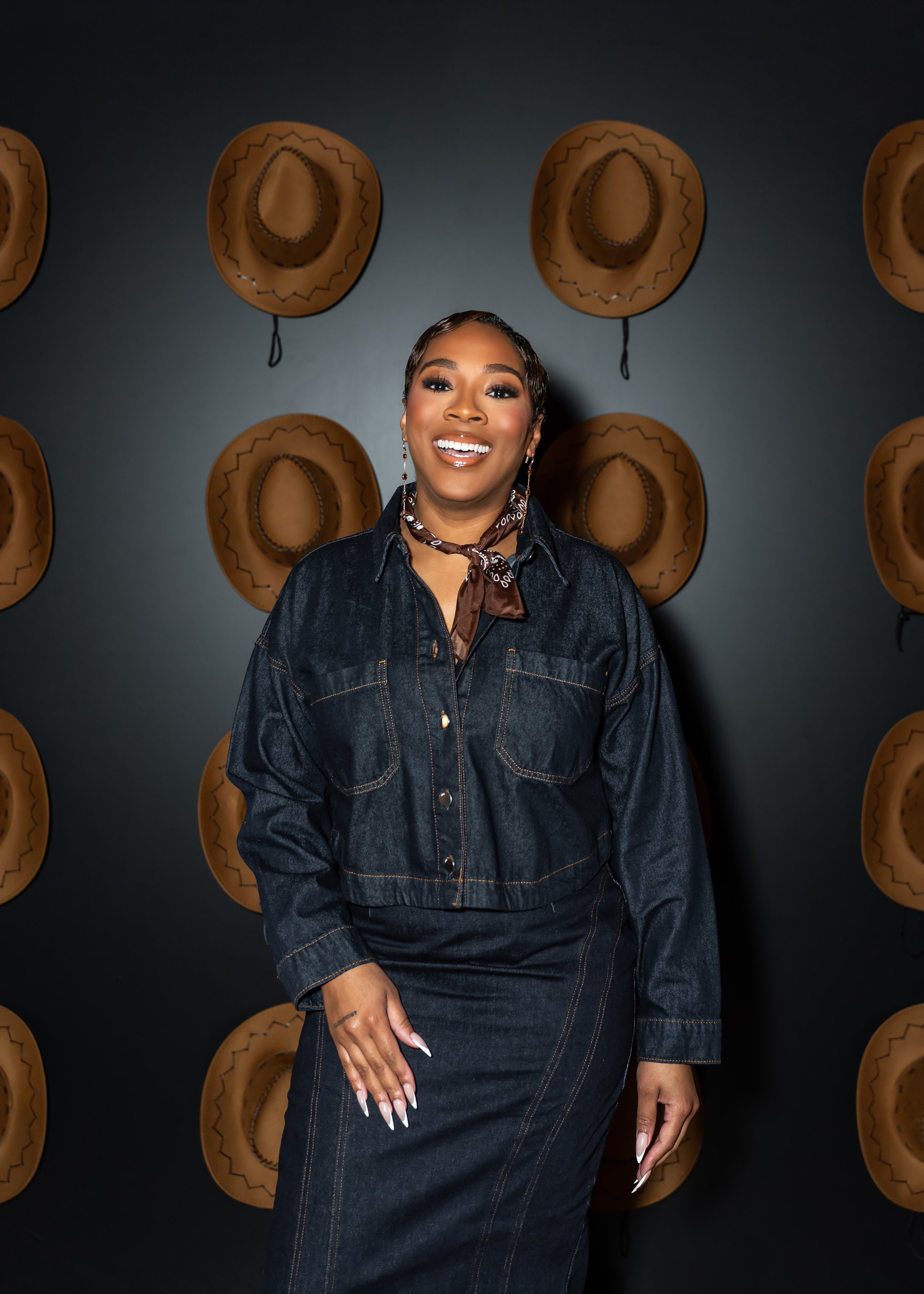 A smiling woman with dark hair styled in waves, wearing a dark denim jacket and matching skirt, standing in front of a dark wall with cowboy hats hanging as decoration.