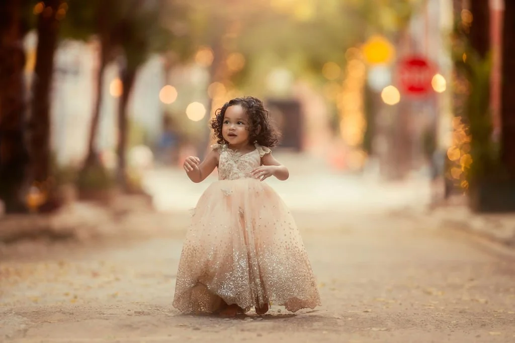Little girl wearing a gold dress standing on Addison Street near Rittenhouse Square, with twinkling lights glowing behind her.