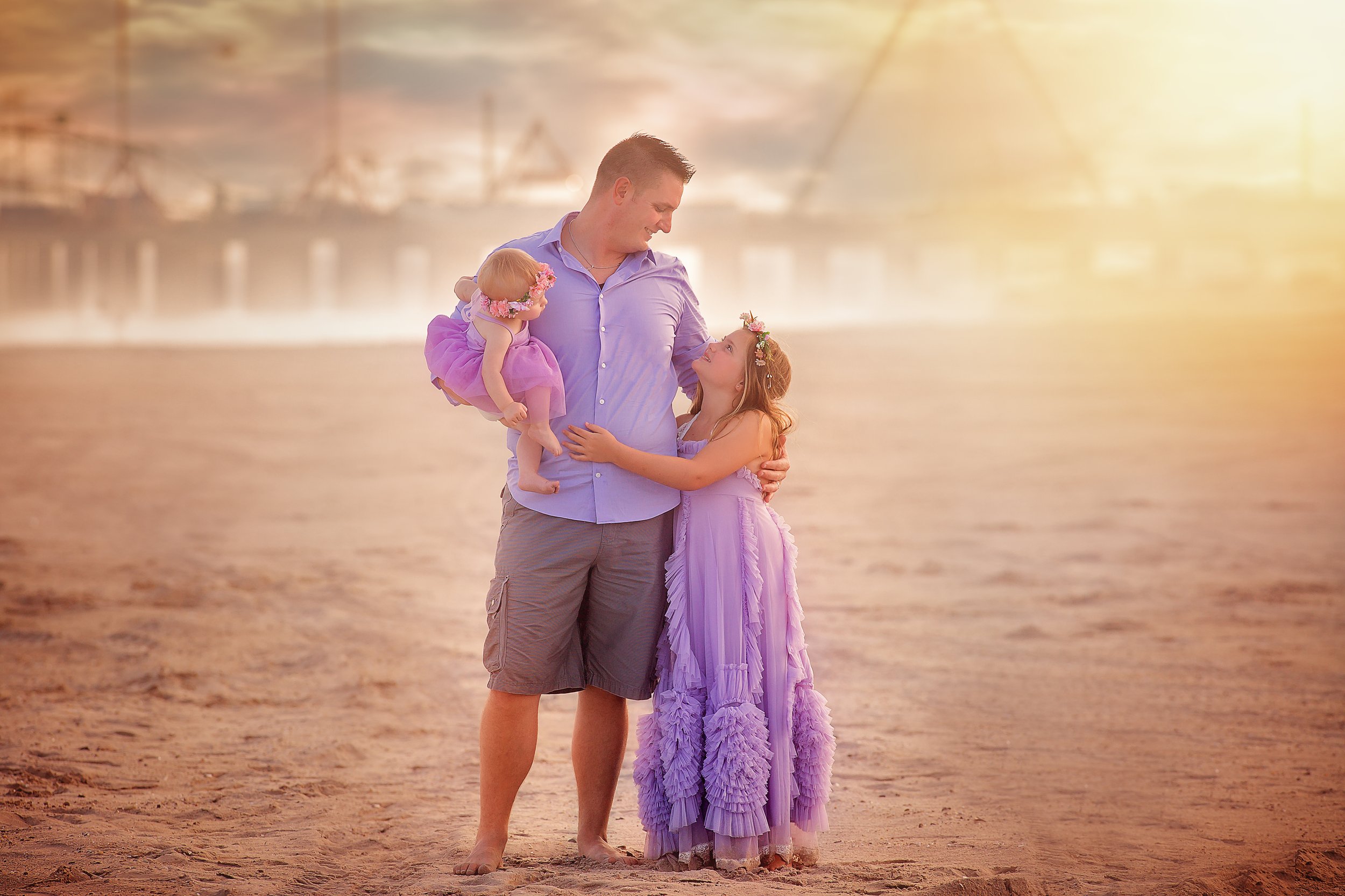 A father standing with his two young daughters during a relaxed family mini session at the beach in soft natural light.