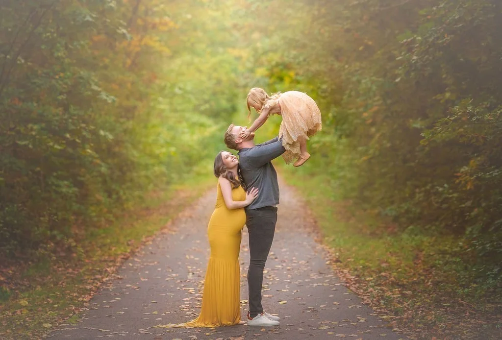 Happy family maternity photo with father lifting daughter and expectant mother smiling during an outdoor portrait session.